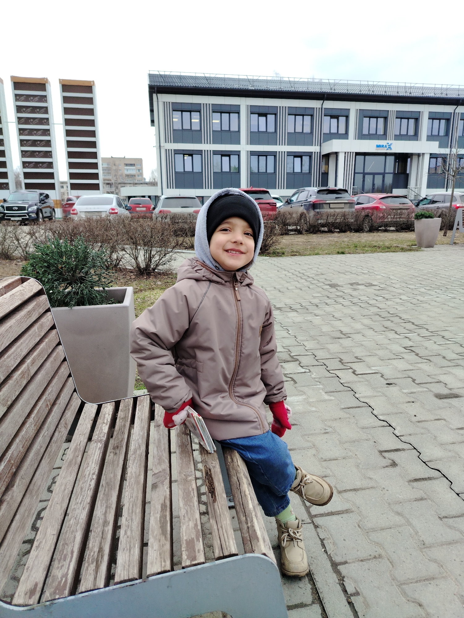 A smiling child, wearing a hooded taupe jacket, dark blue jeans, light tan boots, red gloves, and a dark beanie with a light gray hood pulled up, sits on a wooden bench. The child is looking towards the camera. The bench sits on a light gray brick paved area. Behind the child, there is a modern two-story building with gray and white paneling and dark tinted windows with the text "MIRA X" above the entrance. To the left is a tall, partially visible building with horizontal stripes. A variety of cars are parked in front of the building, separated by landscaping. This scene is in the city of Chaikovsky, Russia.