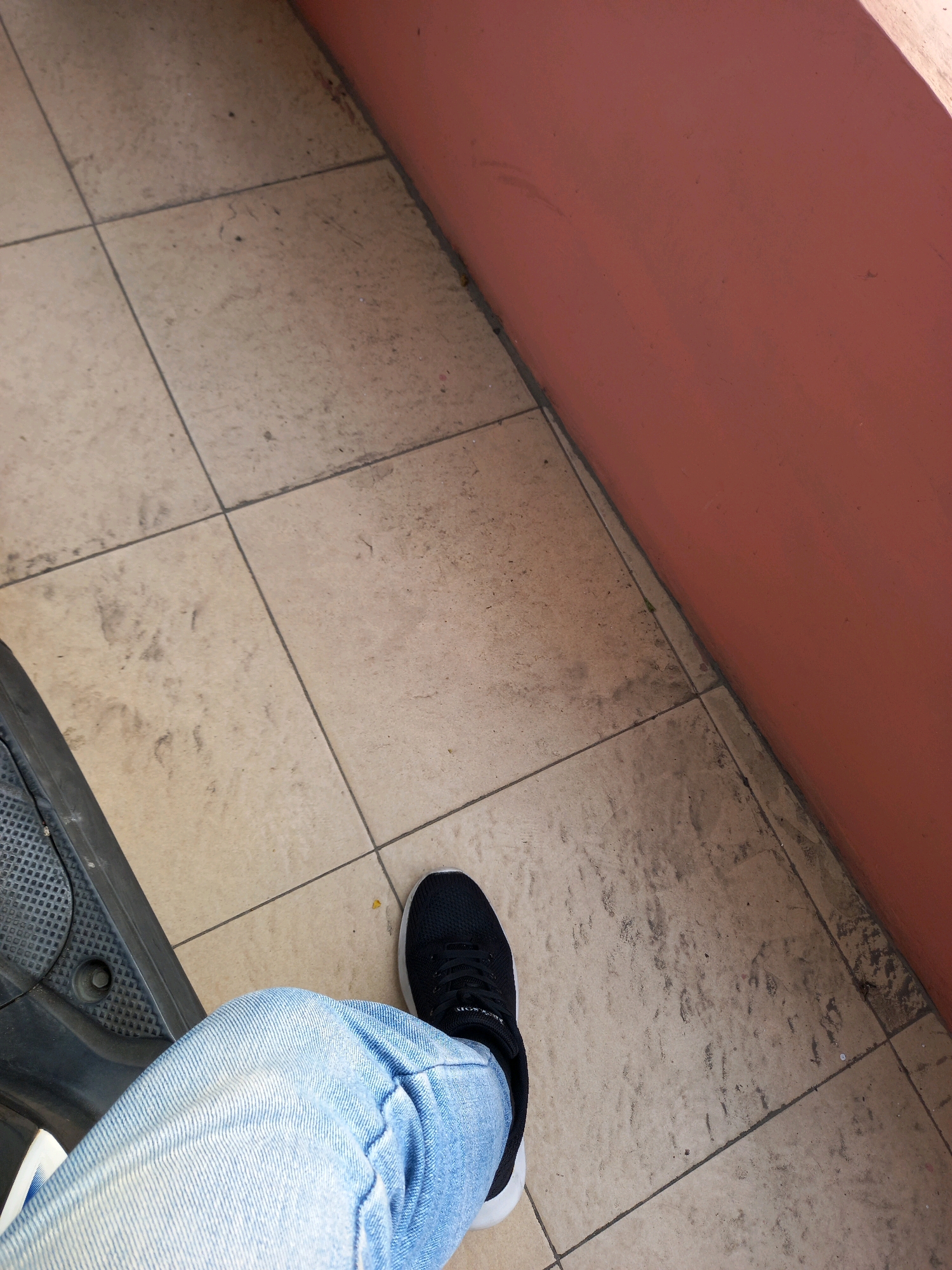The image shows a close-up, angled view of a person's legs and feet, wearing light blue jeans and black athletic shoes, sitting on a dark textured surface, possibly a scooter or motorcycle seat. The background consists of tan, square tiles with dark grout lines, exhibiting some dust and dirt. To the right, a coral-colored wall forms the edge of the scene. The lighting suggests an outdoor setting, possibly during the day. No specific landmarks in Samarinda, Indonesia, are identifiable from this image.