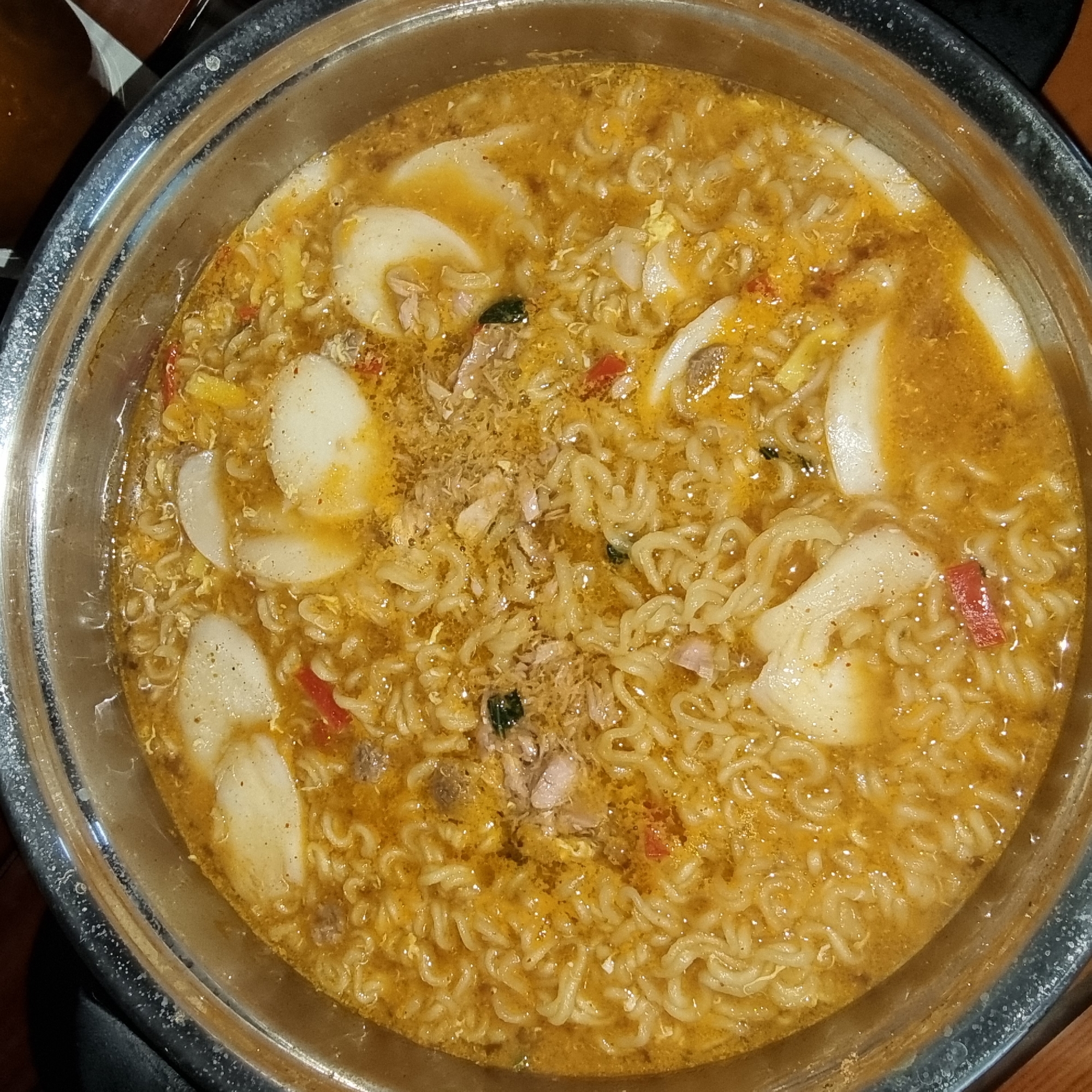 This is a close-up, overhead shot of a steaming pot of ramen with several pieces of tteok (rice cakes) submerged in the broth. The ramen noodles are tightly coiled, and the broth is a rich, golden-orange color, suggesting it's a spicy and savory flavor. Scattered throughout the ramen are small pieces of shredded meat, flecks of what appear to be scrambled egg, and red slivers of chili pepper. The tteok are thick and white, with a slightly uneven surface. The pot itself is made of stainless steel and has a metallic sheen. The background is dark and indistinct, offering no specific environmental details about its location in Dunnae-myeon, South Korea. The image is focused on the food, capturing the texture and appearance of the dish in a rustic, home-style presentation.