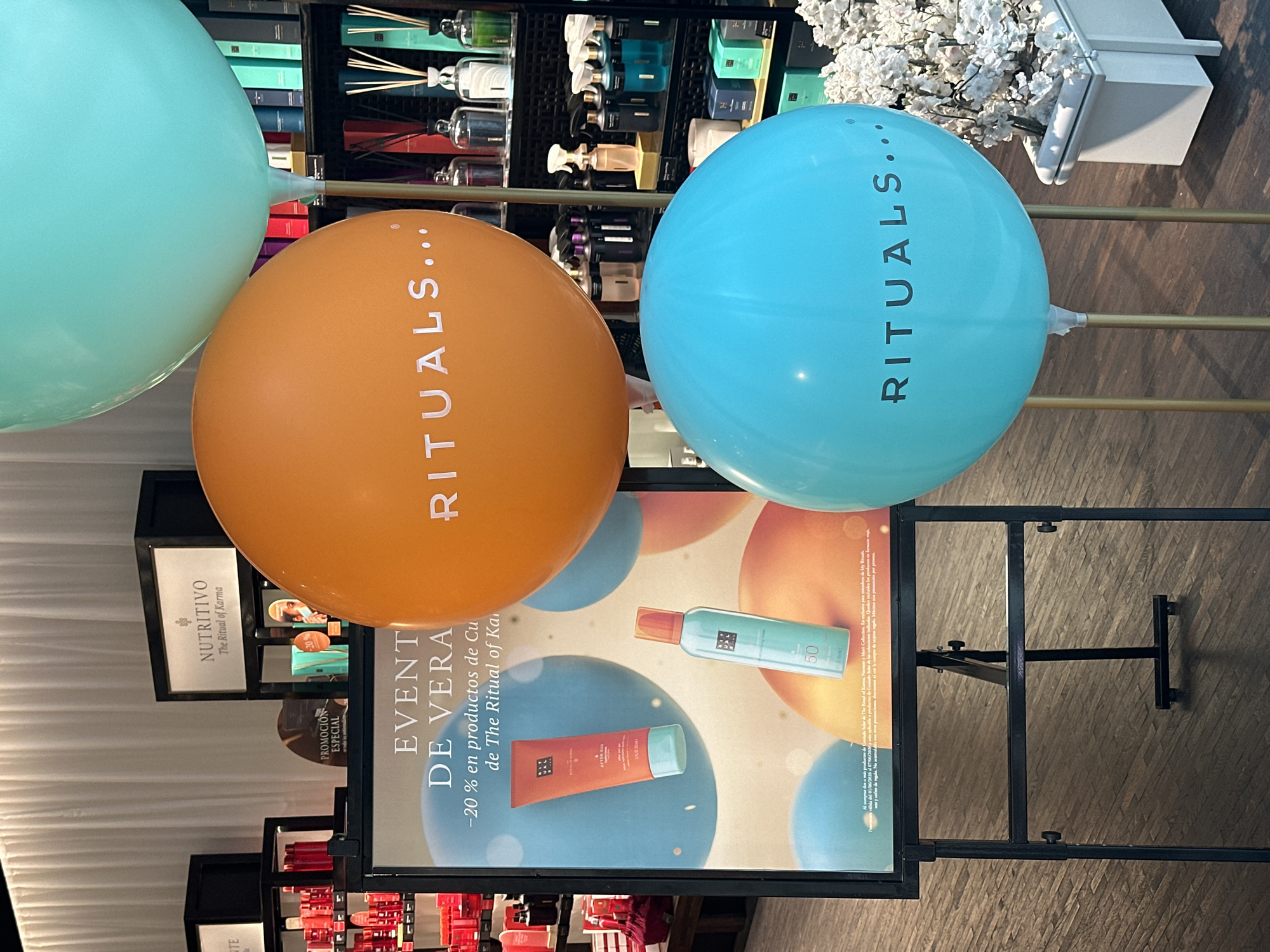 Here is a detailed description of the image:
**General Description**
The image is an indoor shot featuring promotional material for a "Rituals" event. There are several balloons arranged in front of a poster, with shelves of products visible in the background.
**Objects and Subjects**
1. **Balloons:** There are three large, round balloons. One is a light blue color with the word "RITUALS..." printed vertically on it. Another balloon is orange, similarly branded with "RITUALS...".
2. **Promotional Poster:** A poster is visible beneath the balloons, advertising an "EVENTO DE VERANO" (Summer Event) offering -20% off on certain products from "The Ritual of Karma." The poster features images of Rituals products.
3. **Shelves and Products:** In the background, there are shelves stocked with various Rituals products.
4. **Stand:** The poster is held by a black metal stand.
**Scene and Environment**
The scene appears to be inside a store or retail space. The focus is on promoting a special offer, with colorful balloons drawing attention to the promotion. The overall lighting seems bright and artificial, typical of retail environments.
**Overall Impression**
The setup suggests a promotional event aimed at drawing customers' attention to Rituals products, specifically a summer offer from "The Ritual of Karma" line.