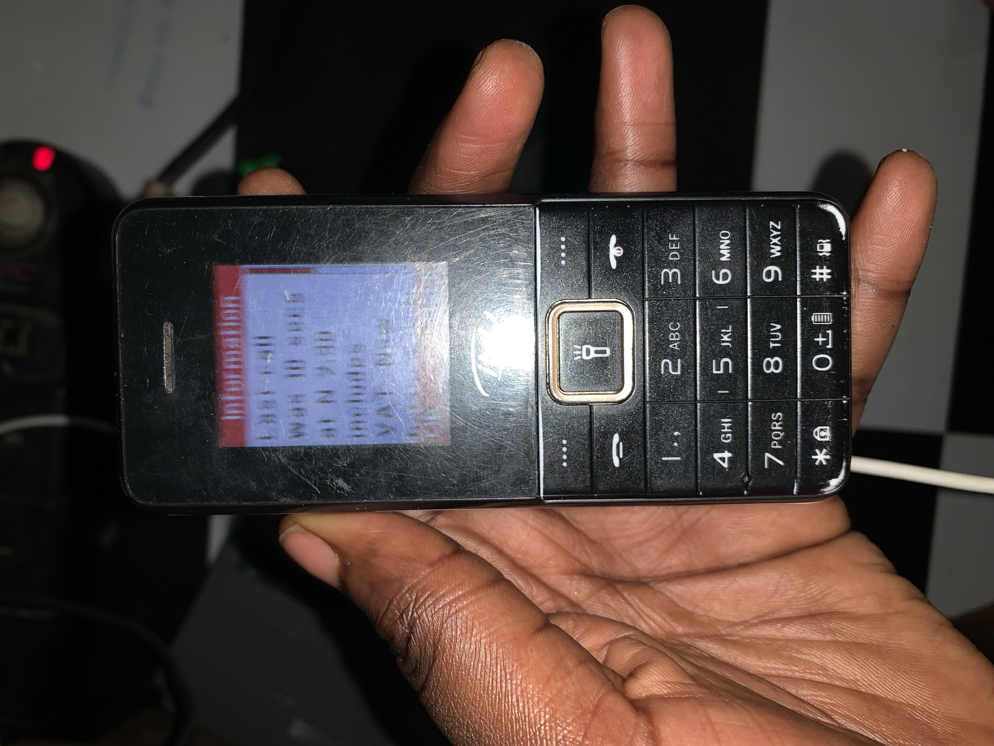 A person is holding a black feature phone in their palm. The phone screen is lit up and displays the word "Information" followed by other text in blue, although the details are difficult to discern due to the image quality. The phone has a physical keypad with alphanumeric keys, and several function keys. The background is dark and partially obscured, but it appears to include a black and white checkered surface. There is no indication of the image being taken in Rafin Par, Nigeria, or any other city in Nigeria.
