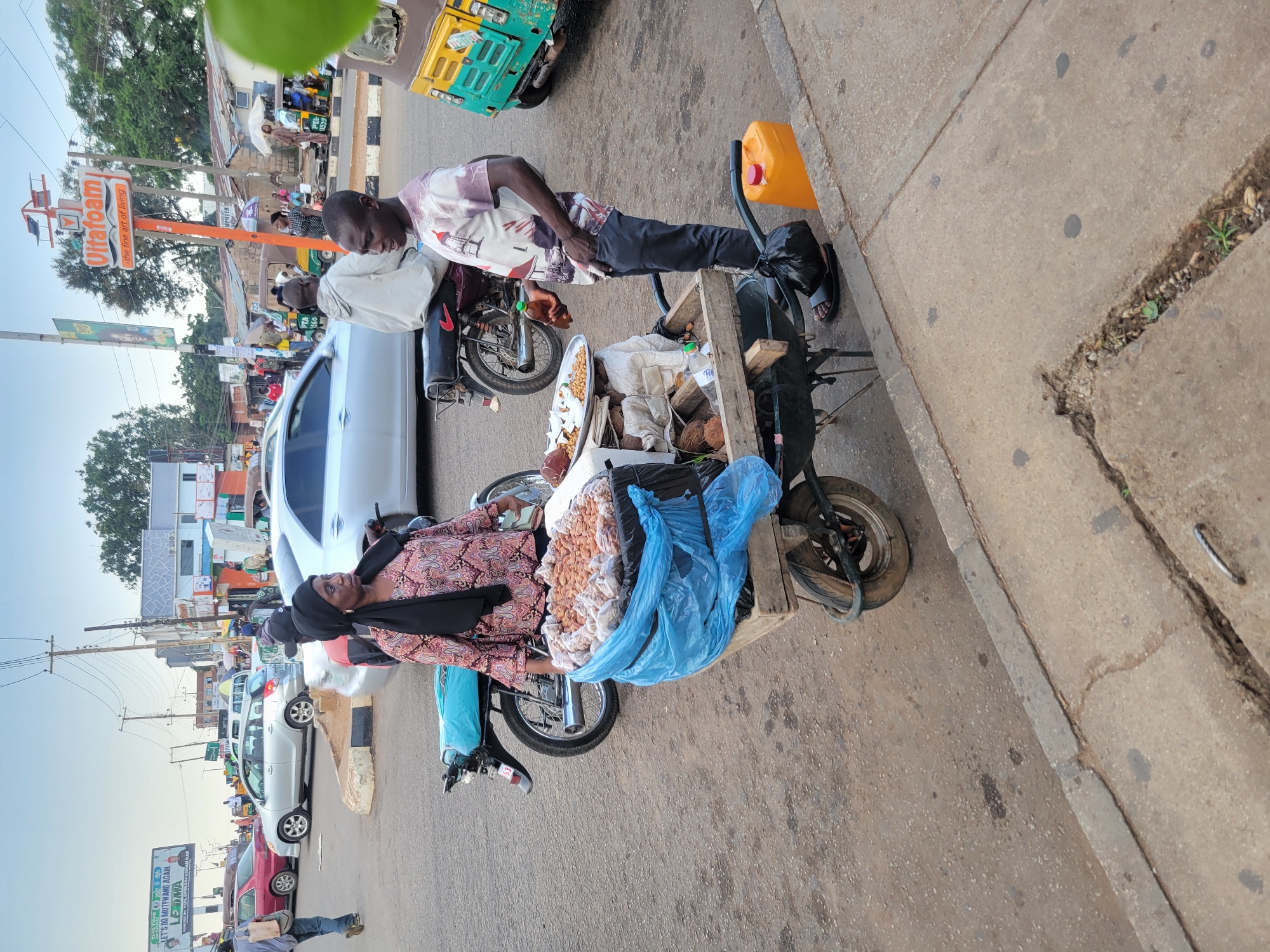 The image depicts an outdoor scene in Barakin Lerawi, Nigeria, likely a busy street intersection. In the foreground, a woman wearing a headscarf and a patterned tunic stands next to a wooden wheelbarrow filled with what appears to be food items, possibly nuts or snacks, contained within blue plastic bags. Beside the wheelbarrow, a man in a patterned t-shirt and dark pants is standing, holding a small bottle and engaging with the woman. Behind them, a motorcycle is parked. Further in the background, a silver car and a red car are visible on the road, along with other vehicles and pedestrians. A prominent "Ultrafoam" sign is visible on a pole. The environment suggests a commercial area with various stalls or shops lining the street, and an overall atmosphere of daily life and commerce.