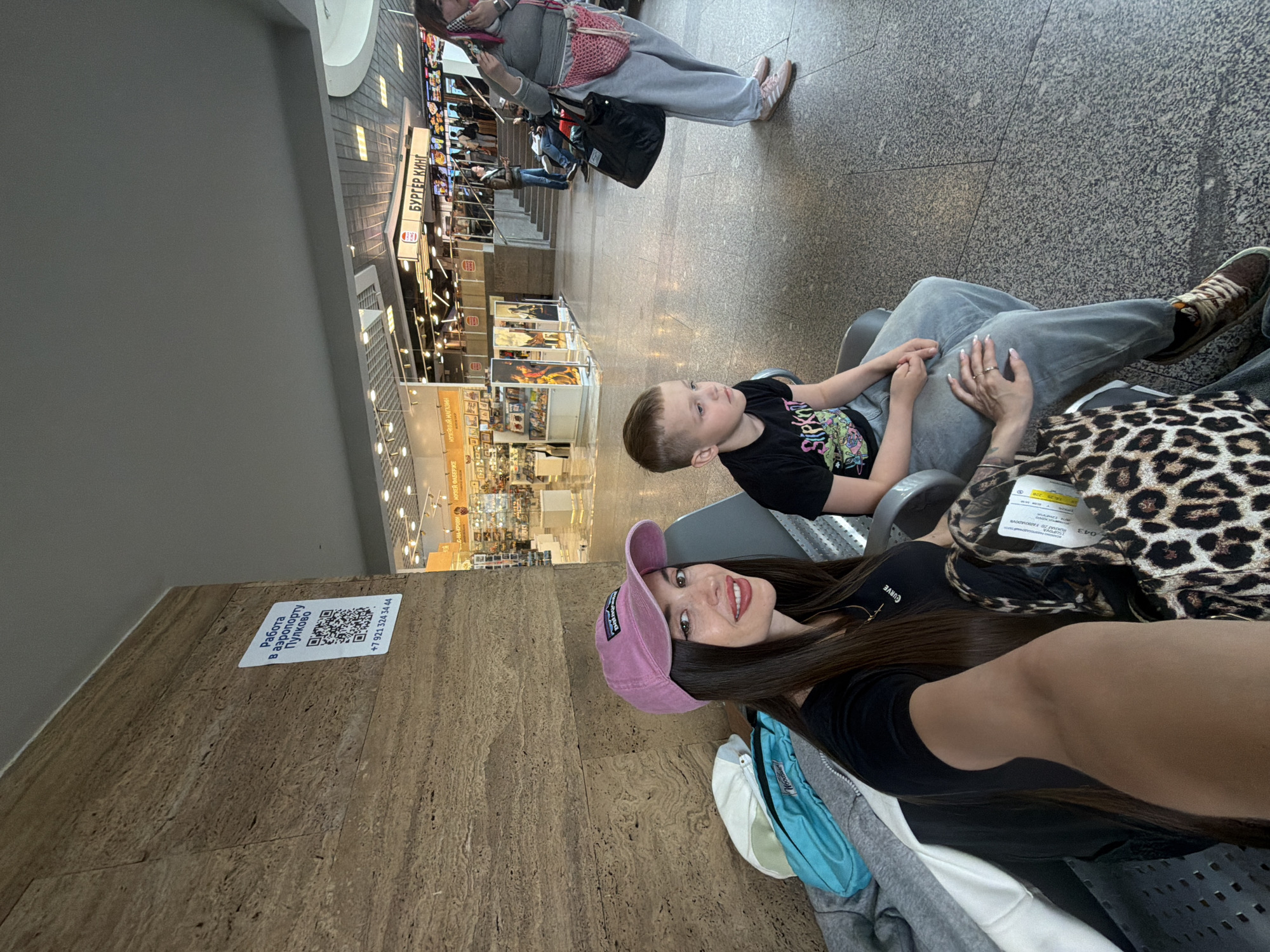 The image captures a selfie of a young woman and a child in what appears to be a waiting area or food court, identified as Pulkovo Airport in Saint Petersburg, Russia. The woman, with long dark hair and a bright smile, wears a black top, a pink baseball cap, and light-colored pants. She is holding hands with a young boy who sits beside her. The boy wears a black t-shirt with a colorful graphic that includes the partially visible text "Slipkn" (likely "Slipknot"), and light-colored pants.
In the background, other people are visible, some standing, some seated. The setting features a tiled floor and walls with a textured, possibly marble, finish. Various commercial establishments line the area, including a prominent "БУРГЕР КИНГ" (Burger King) sign and other signs indicating "МУЗЕЙ ФАБЕРЖЕ" (Faberge Museum) and "МУЗЕЙНЫЙ МАГАЗИН" (Museum Shop). A leopard-print bag rests next to the woman, bearing a tag with "043" visible. The woman's manicured hands, wearing a ring, are clasped with the child's hands.
A sticker on the wall provides a significant location cue: "Работа в аэропорту Пулково" (Work at Pulkovo Airport) along with a QR code and a phone number (+7 921 324 34 44). The woman's top also has the brand name "CURVE" visible. The overall scene suggests a relaxed moment during travel or waiting in an airport terminal.
