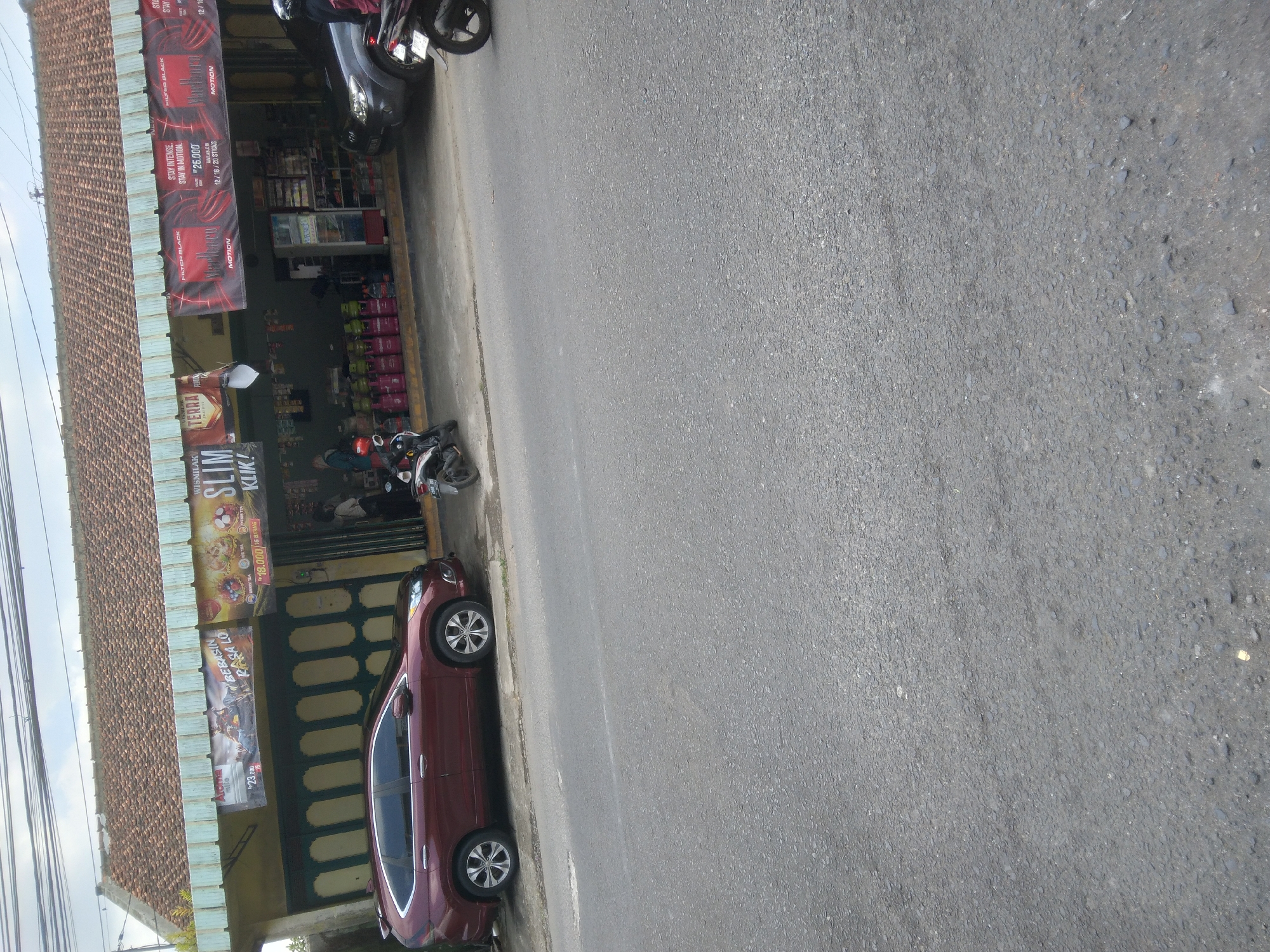 The image depicts a street scene in Purwokerto Wetan, Indonesia, featuring a convenience store with several parked vehicles and gas cylinders outside. A dark red SUV is parked on the left side of the store's frontage, facing towards the right. In front of the store, multiple green and pink propane gas cylinders are neatly arranged in rows on a yellow base. Two motorcycles are parked in front of the store, one dark-colored and the other black. A darker SUV is parked on the right side of the store. The store itself has a covered awning displaying various advertisements, including one for "SLIM KLIK!" and others for Marlboro cigarettes. Two individuals, appearing to be employees or customers, are visible near the store entrance. The street in the foreground is paved with asphalt. The sky above is overcast with some visible power lines. The scene suggests a typical commercial area in Purwokerto Wetan.