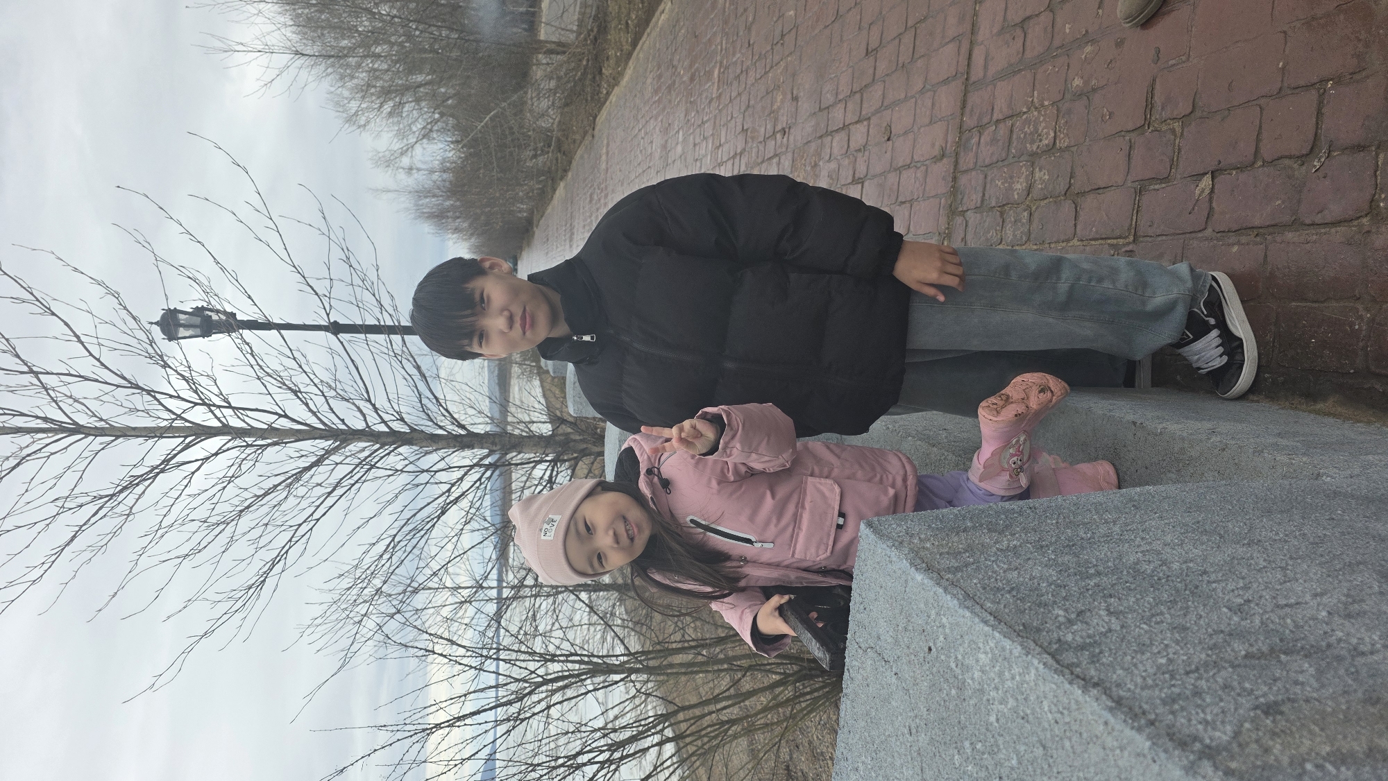 Two children, a boy and a girl, are posing outdoors in Lensk, Russia. The boy is older, wearing a black puffer jacket and jeans, with black and white sneakers. The younger girl wears a pink puffer jacket, a pink beanie, and pink boots with cartoon characters on them. She's holding her hand up in a peace sign. They are near a stone barrier or steps. A bare tree is to the left of the children, and behind the tree is a lamppost and water in the background. Brick pavement is adjacent to the stone barrier, and bare trees can be seen in the distance. The sky is overcast.