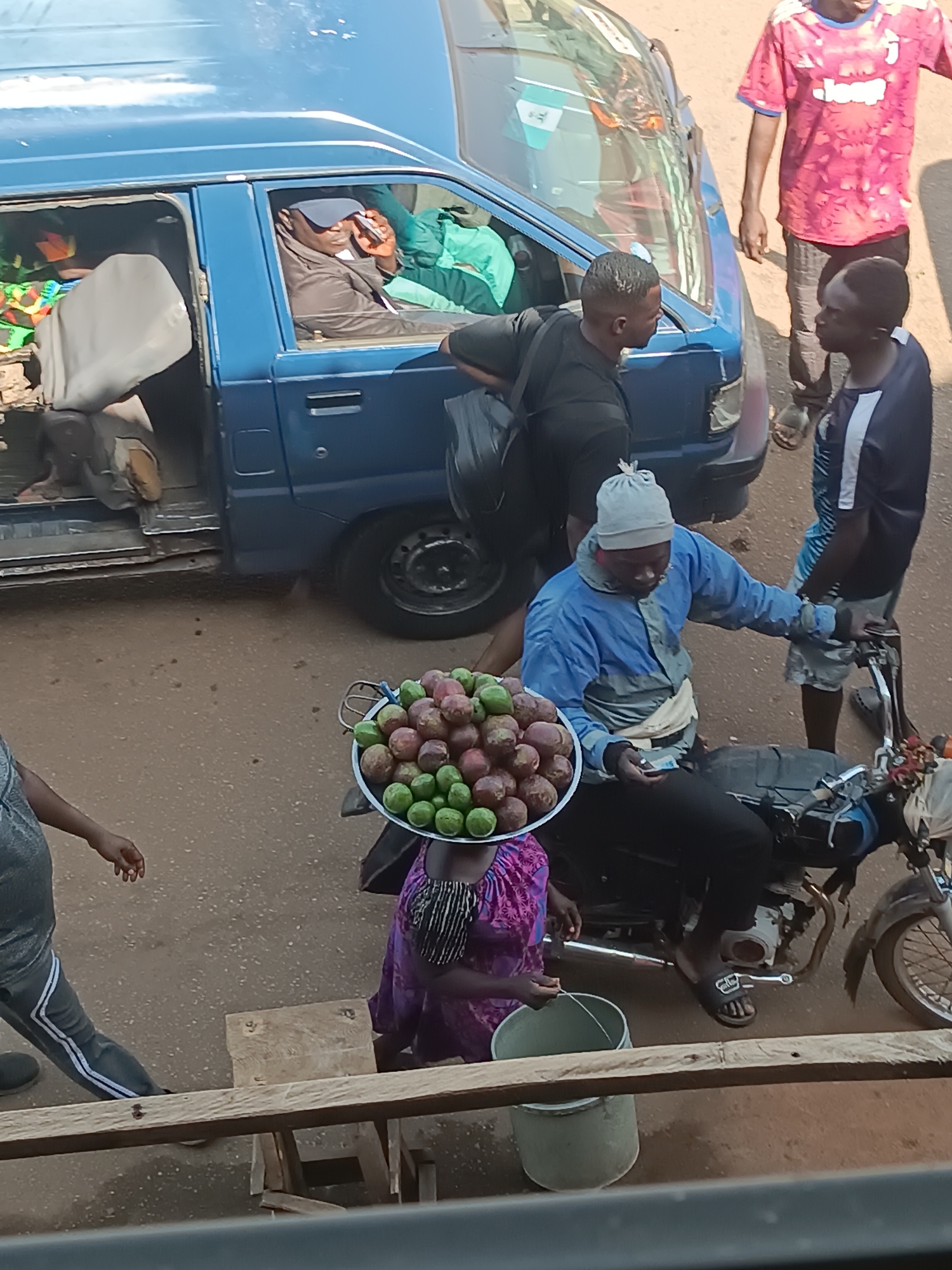 The image depicts a bustling street scene in Bukuru, Nigeria. A blue van with its door open is visible, with two individuals inside, one of whom is on the phone. A man with a black bag stands outside the van, talking to another man. A motorcycle is parked nearby, with a vendor seated on it wearing a blue jacket and a hat. He appears to be handling money. On the vendor’s head is a tray loaded with green and purple fruit. A woman in a purple dress stands beside the motorcycle holding a small bucket. Another person is partially visible on the left side of the frame, walking away. A wooden structure is visible in the foreground, possibly a railing. The background shows a man wearing a pink shirt with blue sleeves. The road surface is brown and dusty.