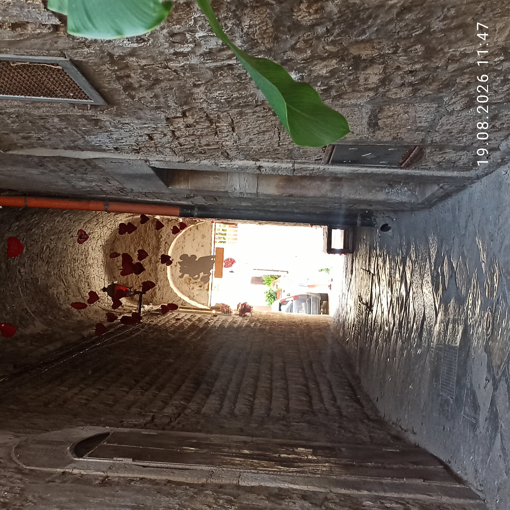 This image depicts a narrow, arched alleyway in Ruvo di Puglia, Italy, decorated with numerous red heart-shaped ornaments suspended from the ceiling. The walls of the alley are constructed from rough, textured stone blocks. Sunlight streams in from the far end of the alley, illuminating the cobblestone pavement. At the end of the alley, a person in a white top and shorts is visible near a car. A green leaf from a plant is positioned in the foreground on the right side of the image. The date and time stamp "19.08.2026 11:47" is overlaid in the lower right corner.