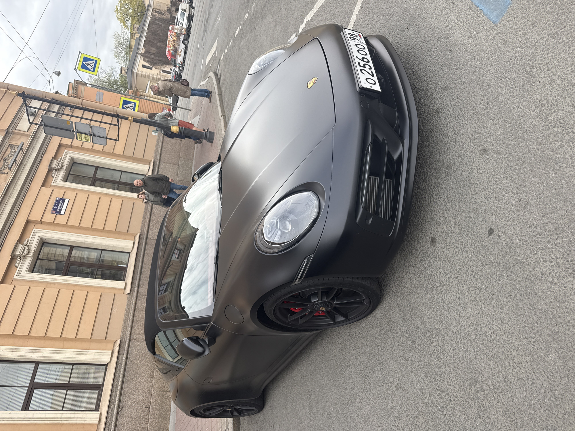 The image depicts a matte black Porsche sports car parked on a city street. Pedestrians are visible in the background, alongside buildings with classic architectural details. Road markings and street signs are present. The car's license plate reads "O 256 ОО 98". The setting is likely within the city of Saint Petersburg, Russia.