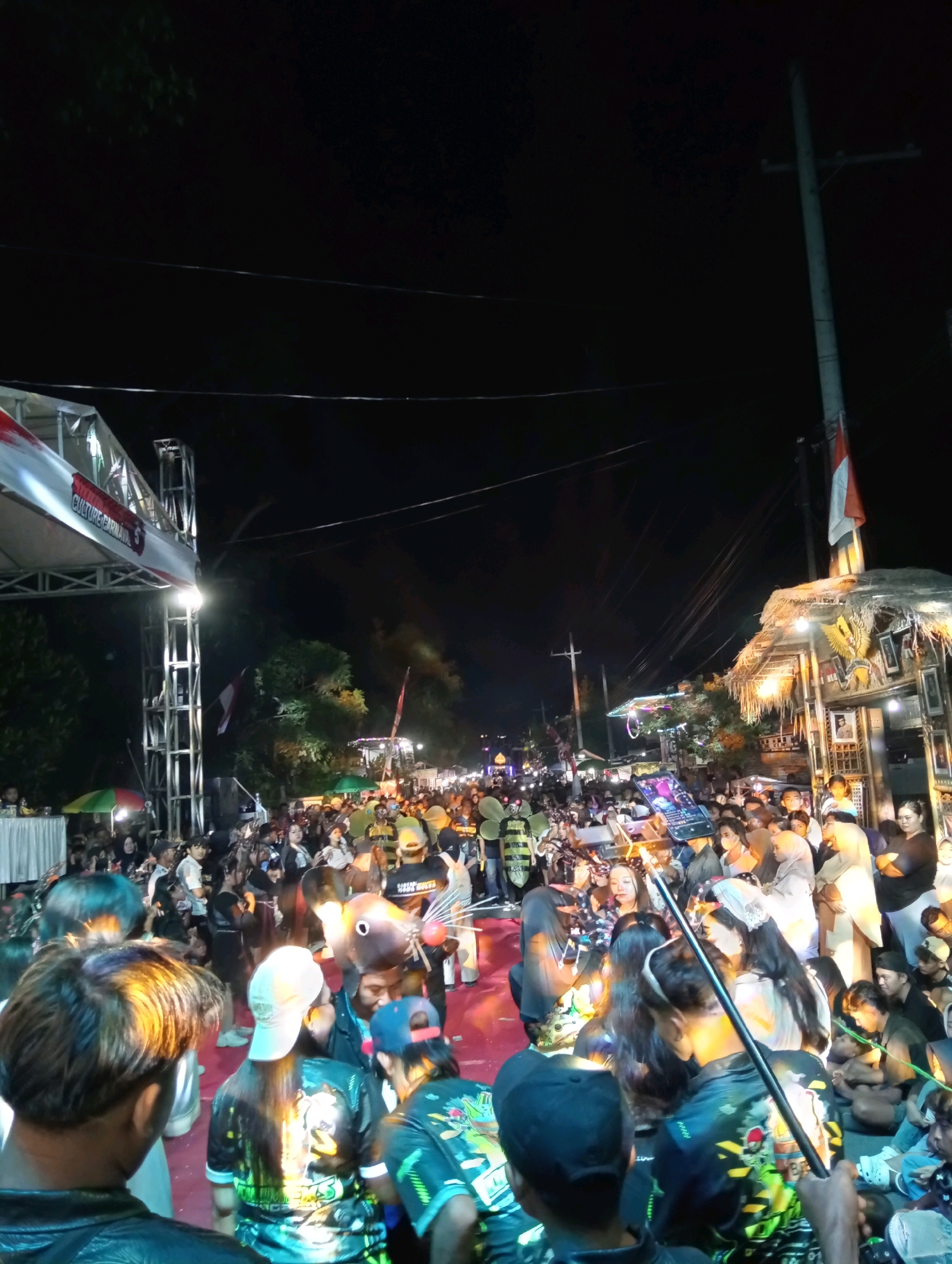This is a nighttime street scene in Curah Malang, Indonesia, likely during a cultural event or carnival. A large crowd of people are gathered on a street illuminated by stage lights and string lights. In the foreground, the backs of several individuals are visible, some wearing brightly colored shirts. In the middle ground, a stage structure with banners is set up on the left side of the image, and to the right, a thatched-roof building with Indonesian flags and portraits is visible. People in the crowd are wearing various clothing, some in costumes, including one person in a large, fish-like headpiece. Some individuals are holding cell phones aloft. The overall atmosphere appears festive and crowded.