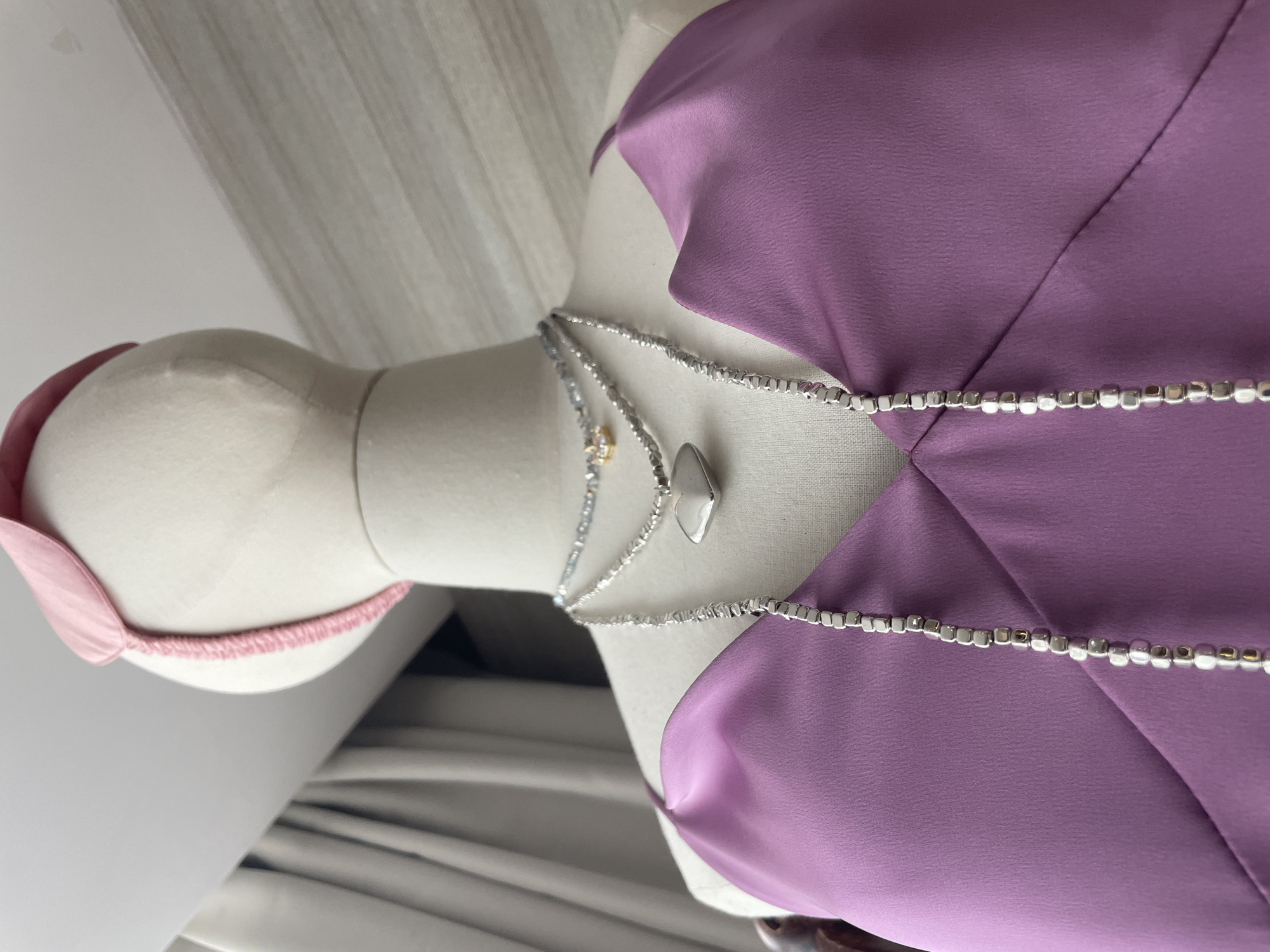 A mannequin bust is draped in a purple satin top. Three necklaces are layered on the mannequin's neck. The top necklace appears to be a fine chain with small beads and a star-shaped charm. The middle necklace features a faceted, heart-shaped pendant. The bottom necklace is a thicker chain with square silver beads. The background consists of a blurred curtain and a wall with horizontal lines. The scene is indoors, and no specific location in Bac Nha Trang Ward, Vietnam can be identified from the image.