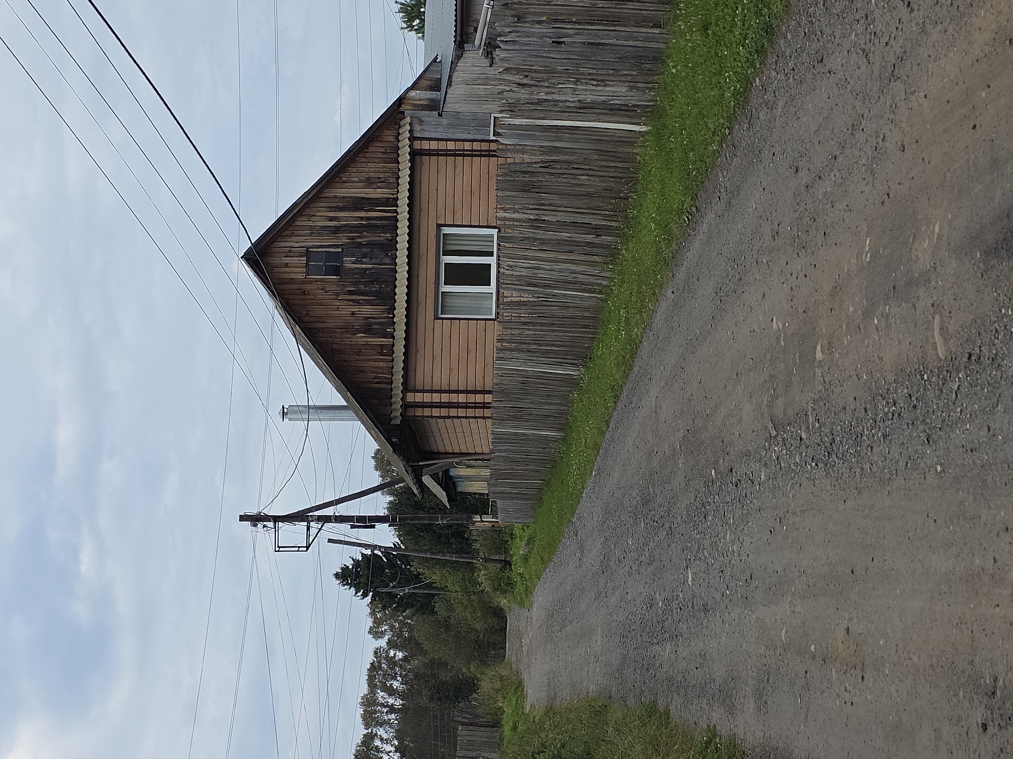 The image depicts a rural scene on an overcast day, likely in or around Magnitka, Russia. A gravel road curves from the foreground into the mid-ground, bordered by grassy verges and a weathered wooden fence. To the right of the road stands a wooden house with orange siding and a steeply pitched, darker wood roof. A single window with white curtains is visible on the lower floor. Above this, on the gable end, is a small, dark square window. A metal chimney extends from the roof. A wooden utility pole with electrical wires stretches across the sky above the house and road. Trees line the left side of the road further into the distance. The overall impression is one of a quiet, somewhat rustic, residential area.
