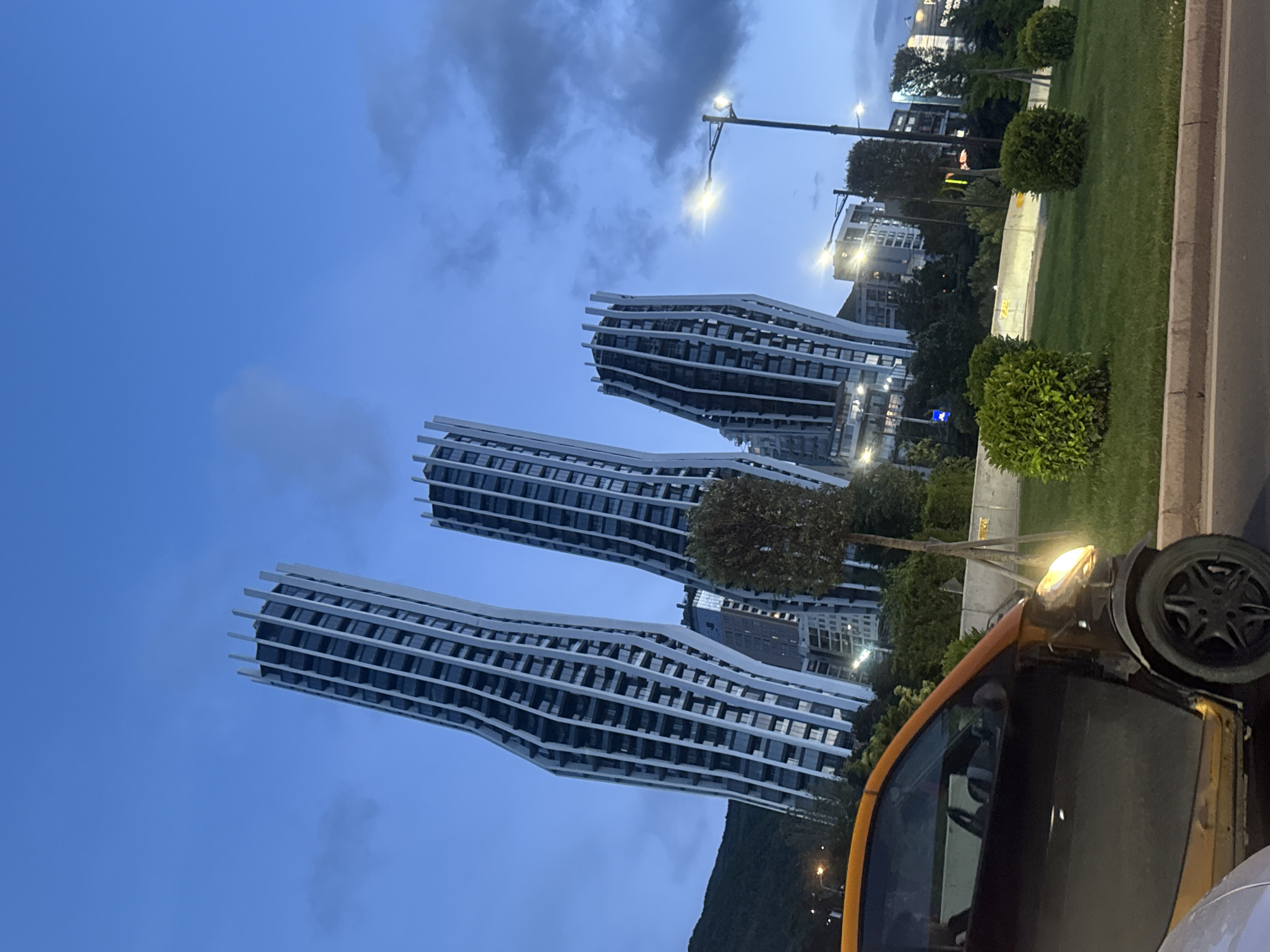 The image shows three modern, high-rise apartment buildings with undulating facades in Tbilisi, Georgia. The buildings are primarily white with dark windows. The sky is a dusky blue with some cloud cover. A portion of a yellow car is visible in the foreground, partially obscuring the view. There is a green lawn and trees bordering a street to the right of the buildings. In the background, additional buildings and street lights are visible. The scene appears to be taken in the evening.