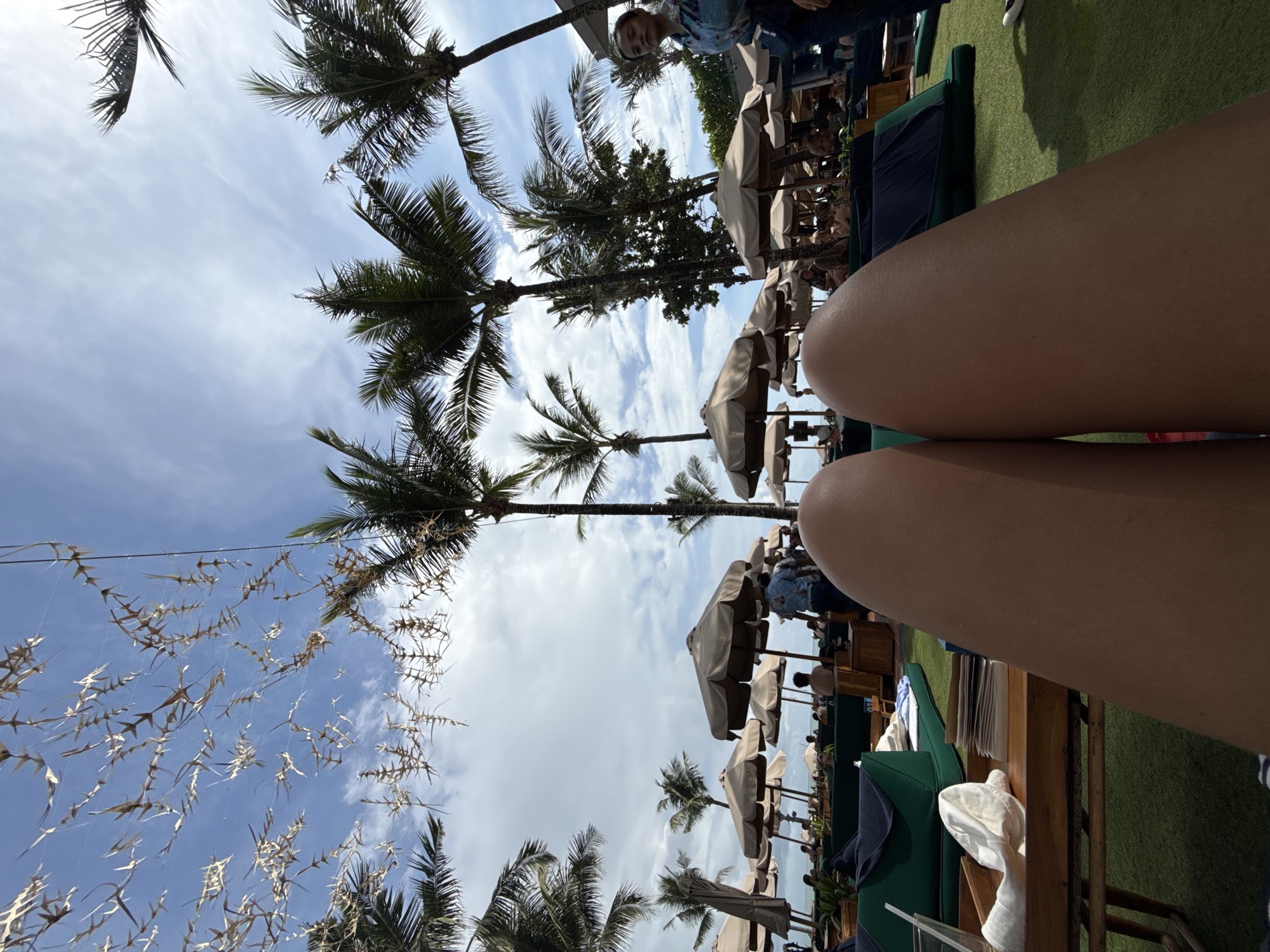 The image, taken in North Kuta, Indonesia, depicts a tropical beach scene. Palm trees are visible against a sky with scattered clouds. Umbrellas provide shade over what appears to be beach chairs or loungers where people are sitting. The legs of someone are in the foreground, while the background features green artificial grass. Hanging decorations create an interesting visual detail.