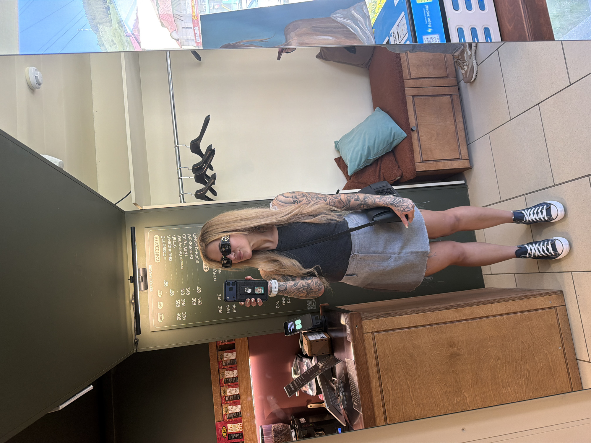 The image captures a woman taking a selfie in what appears to be a cafe in Gorno-Altaysk, Russia, according to the visible text "Бері заряд!" on a nearby box. The woman has blonde hair, sunglasses, tattoos on her arms, and is wearing a black top and a grey skirt. She is holding a black phone with stickers on the case.
Behind her is a menu board written in Russian, suggesting the cafe serves coffee. There is a wooden counter with a coffee machine and other items related to coffee preparation. The cafe is reflected in a mirror, showing additional seating with a blue cushion. The floor is tiled. Through the mirror's reflection, a building or structure can be seen outside, possibly indicating an urban setting. The woman is standing next to a dark green wall. She is wearing black and white sneakers.