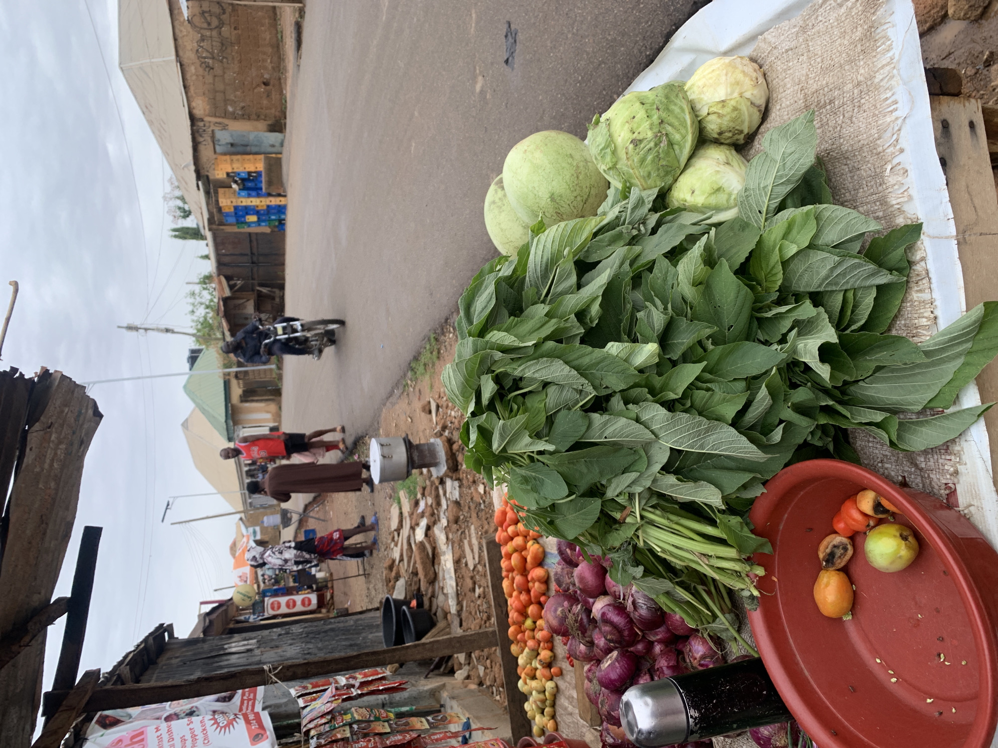 The image depicts a vegetable stand in Barakin Lerawi, Nigeria. On the stand, there are cabbages, leafy green vegetables, tomatoes, and red onions. A red plastic bowl containing what appear to be spoiled vegetables sits on the corner of the table. In the background, pedestrians walk along the roadside and a motorcyclist rides down the road. Crates of beverages are stacked against a building, indicating a commercial area. The stand is adjacent to a weathered structure with visible signs of wear and tear.