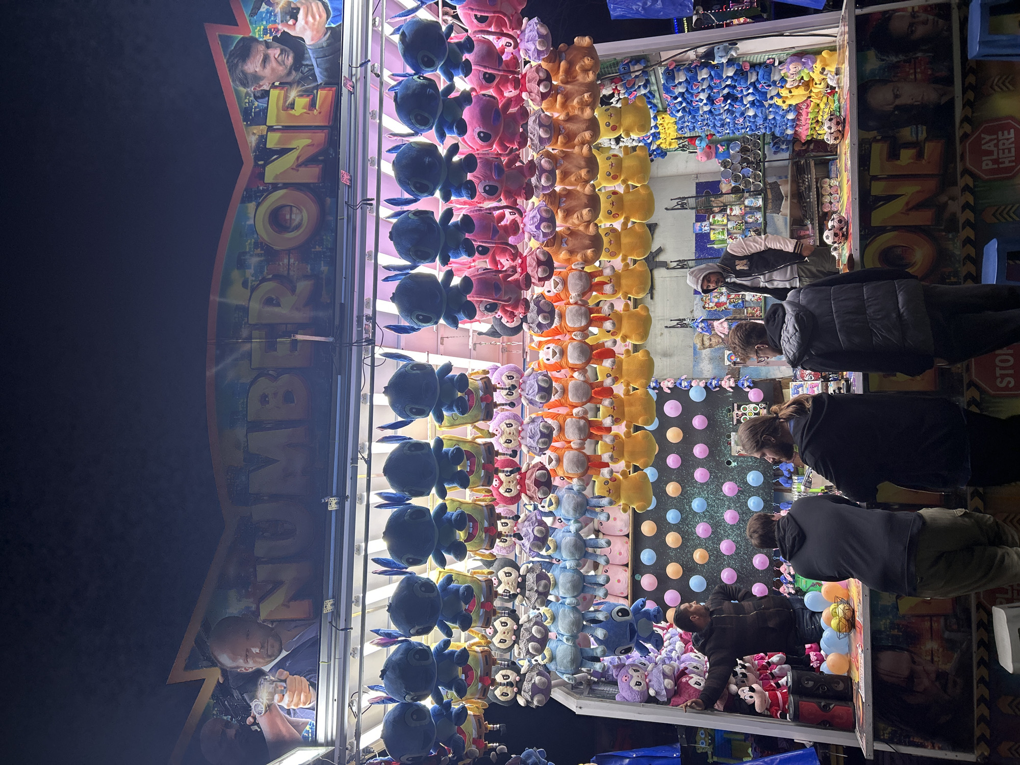 The image depicts a carnival-style game booth, likely located in Plovdiv, Bulgaria during a public fair or festival. Rows of stuffed animal prizes are arranged vertically along the back wall of the booth, including blue, pink, orange, and yellow characters. Several people, mainly adults, are standing at the booth, observing or participating in the game. A section of the booth displays colorful balloons. A sign reading "NUMBER ONE" is visible on both sides of the booth, with images of men holding weapons. The lighting suggests it is evening or nighttime.