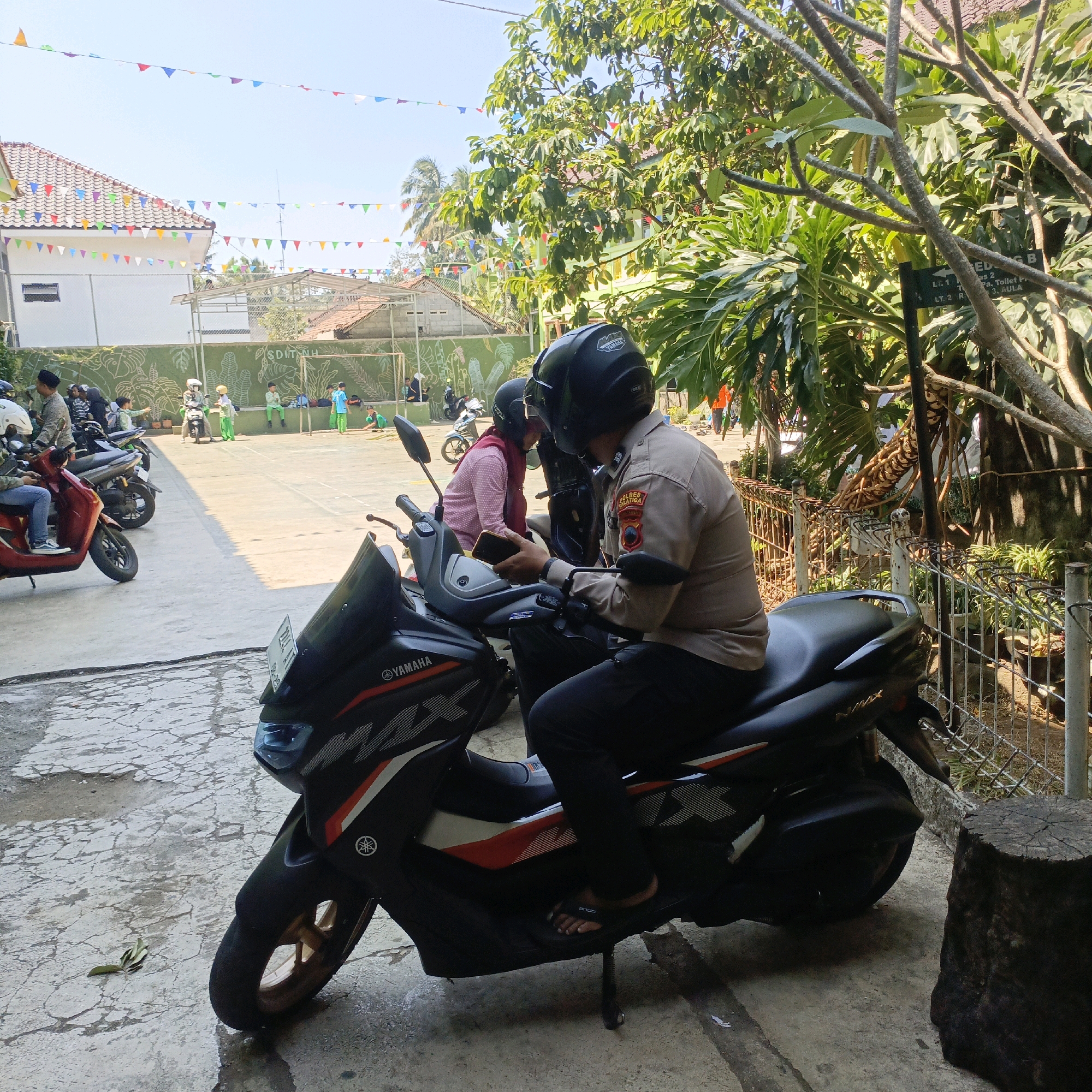 The image depicts an outdoor scene on a sunny day, likely within the grounds of an educational institution in Salatiga, Indonesia. In the foreground, a person wearing a police uniform, complete with a helmet and badge, is seated on a black Yamaha NMAX motorcycle. The officer is looking at and holding a smartphone. Another person wearing a maroon headscarf and a helmet is seated on a second motorcycle to the left of the officer.
The background reveals a courtyard or field area where children in school uniforms, many wearing helmets, are present. Some children are playing or standing in groups, while others are near motorcycles. A green wall with a mural of plants and the text "SD IT AN NAH" is visible behind the children, indicating the name of the school. Houses with red-tiled roofs and colorful triangular flags strung across the sky add to the festive or event-like atmosphere. Trees with lush green foliage and some bare branches are present, along with a metal fence and a wooden tree stump in the right foreground. The ground is paved with concrete, showing signs of wetness and cracks.