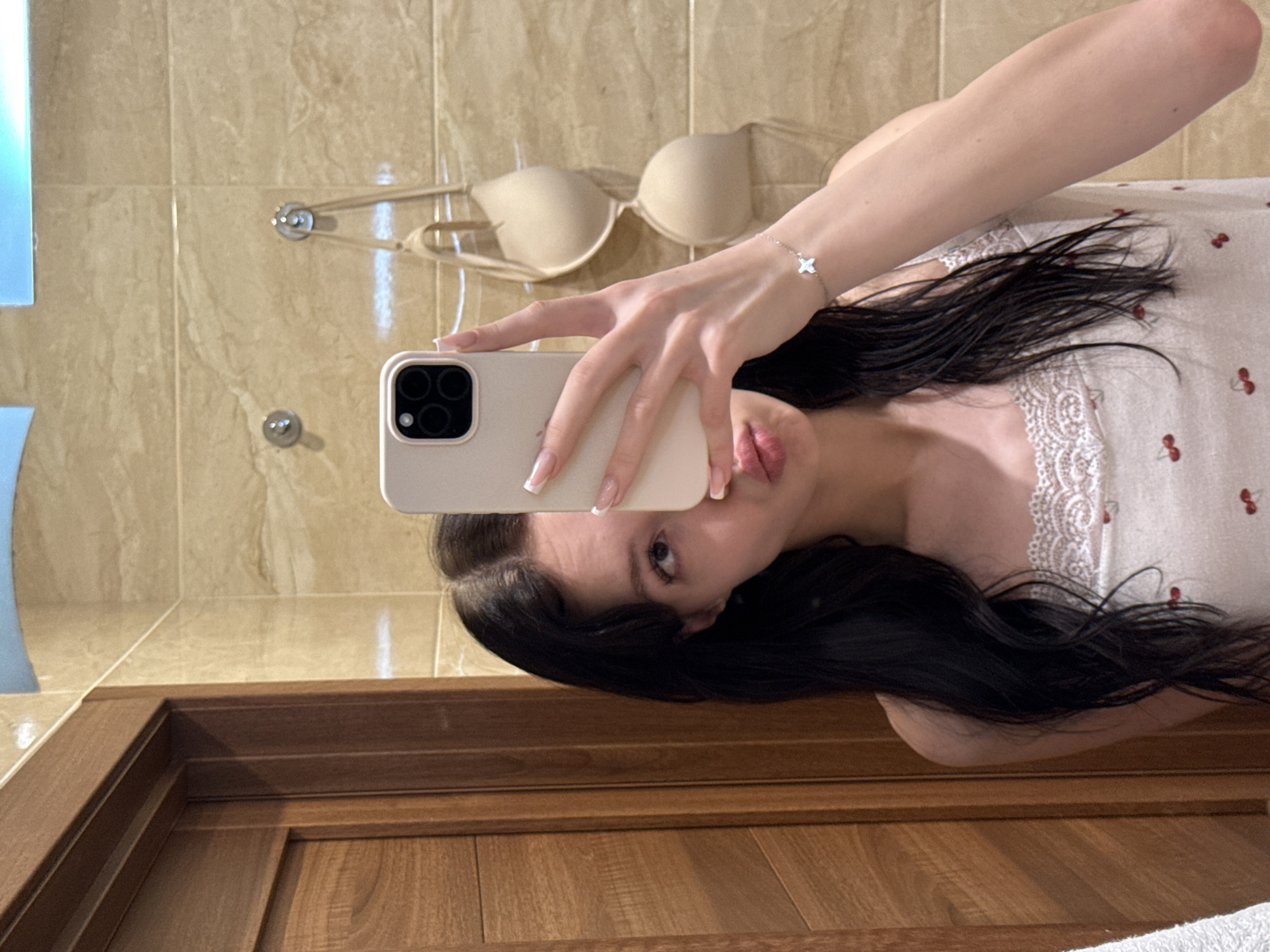 In an indoor bathroom environment, a young woman with long, dark hair is taking a mirror selfie. She is wearing a camisole with a lace trim and a cherry pattern. Her left hand is holding up a smartphone to capture the reflection. A beige bra is hanging on the wall behind her, and the walls are tiled with a marble-like pattern. A wooden door is visible on the left side of the frame. The overall impression is a casual, personal moment captured in a domestic setting. The location is identified as being in the city of Khimki, Russia.