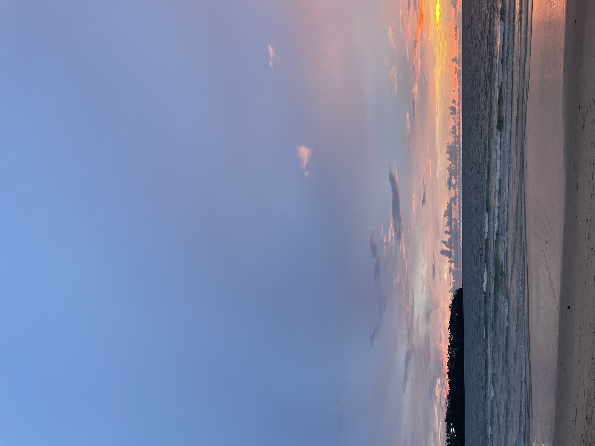 The image depicts a serene beach scene at sunset in Choeng Thale, Thailand. The sky is a gradient of blues, pinks, and oranges, with scattered clouds catching the last rays of the sun. A calm, deep blue ocean stretches across the mid-ground, with gentle waves lapping at the sandy shore. The water reflects the warm colors of the sunset. In the distance, a lush, dark green headland juts out into the sea, dotted with buildings that are barely discernible. The foreground is a sandy beach, wet in places near the water's edge, showing subtle reflections. The scene is devoid of people and any specific objects, focusing solely on the natural beauty of the coastal environment at twilight.