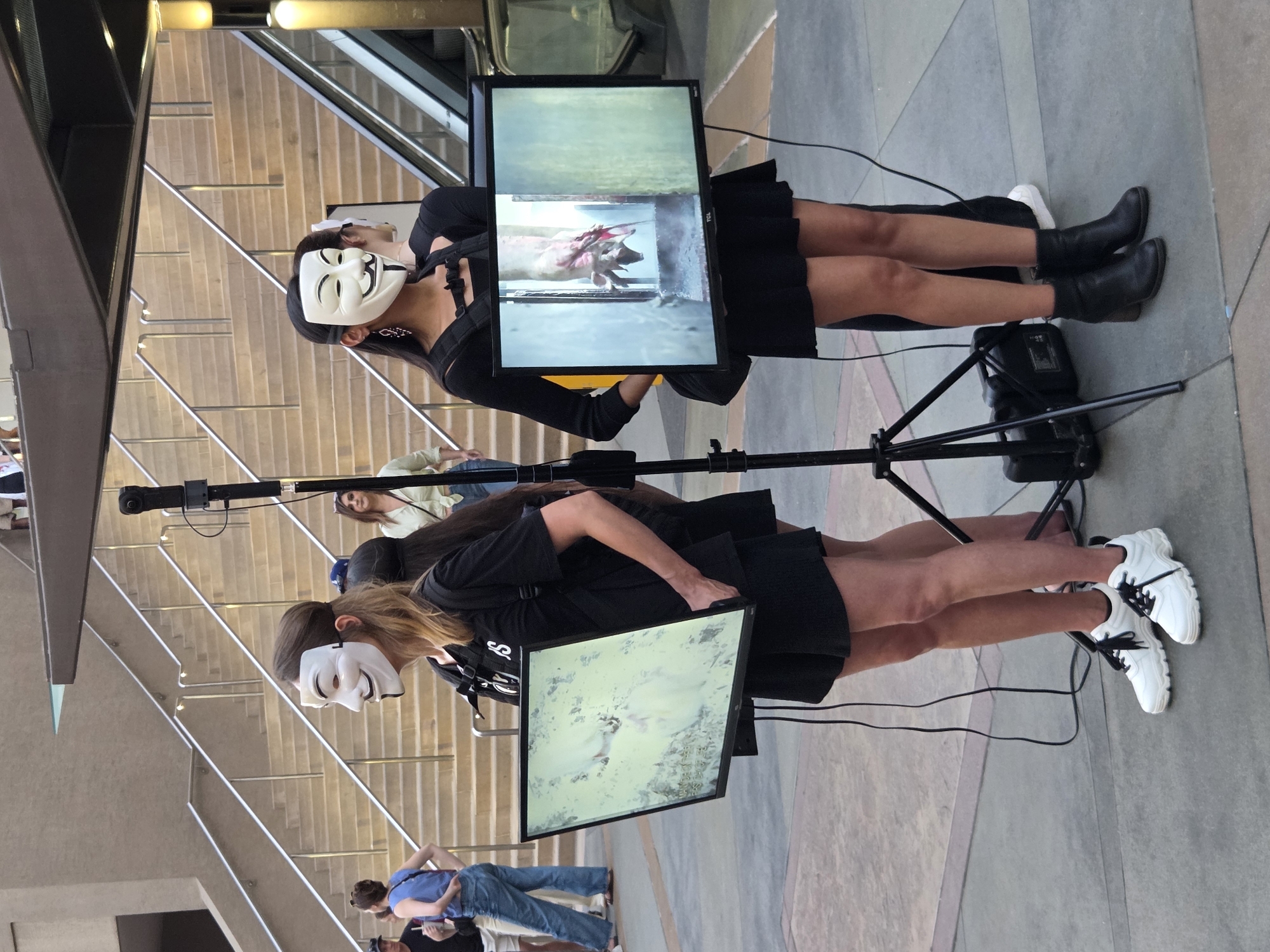 In Burbank, California, at street level, two individuals wearing Guy Fawkes masks are standing side-by-side. Both are dressed in black, with one person wearing shorts and white sneakers, and the other wearing a skirt and black boots. Each is holding a flat-screen monitor, displaying images on their screens. A tripod with a camera is positioned between them, and a black speaker is visible on the ground. In the background, a set of concrete stairs with metal railings leads upwards, and an escalator is visible to the right. Several other people are present in the scene, some walking and others standing, with a woman in a light-colored shirt visible near the stairs. The overall environment suggests an outdoor public space.