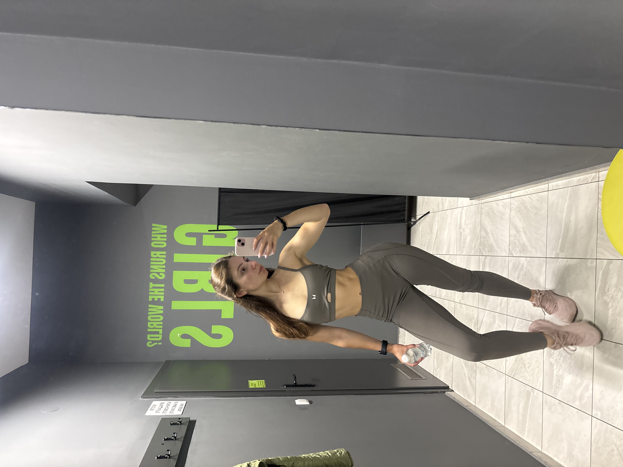 In a modern gym locker room with gray walls and tiled floors, a young woman with long brown hair takes a mirror selfie. She is wearing a grey sports bra and matching leggings, and pink athletic shoes. In her left hand, she holds a clear plastic water bottle. A black smartwatch is visible on her left wrist. The wall behind her features bright green text, partially obscured by her reflection, which reads "WHO RUNS THE WORLD? GIRLS". A coat rack with a green jacket is mounted on the left wall. The overall environment suggests a clean and functional fitness space. The image does not contain information indicating the specific location within Varna, Bulgaria.