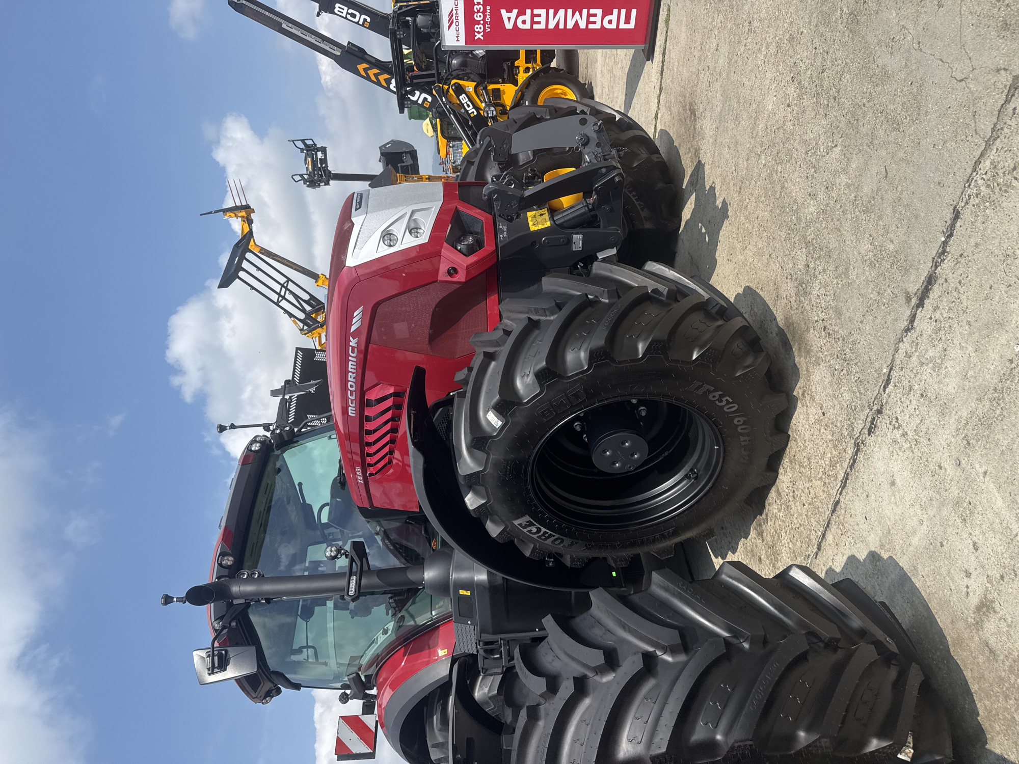 This image captures a vibrant display of agricultural machinery, likely at an outdoor exhibition or dealership in Stara Zagora, Bulgaria. The central subject is a striking red and silver McCormick X8.631 VT-Drive tractor, positioned prominently on a concrete surface. The tractor is equipped with large, rugged tires, showing markings such as "IF650/60R38" and "540/65R30 RADIAL," indicating its heavy-duty capability. Its front end, headlights, and part of the exhaust pipe are clearly visible, showcasing its modern design.
In the background, several yellow JCB machines are partially visible. One appears to be a telehandler or loader, with its boom extended upwards, fitted with a bale spear or fork attachment. A red vertical sign stands to the right, bearing the Cyrillic word "ПРЕМИЕРА" (PREMIERE) in large white letters, along with "MCCORMICK X8.631 VT-Drive," suggesting the tractor is a new model being introduced. The surrounding ground is paved with concrete, exhibiting some cracks, and the setting is under a bright, partly cloudy sky, indicating daytime. The clear light casts shadows on the ground beneath the machinery. No people are visible in the scene, which focuses entirely on the impressive scale and modern design of the heavy-duty equipment.