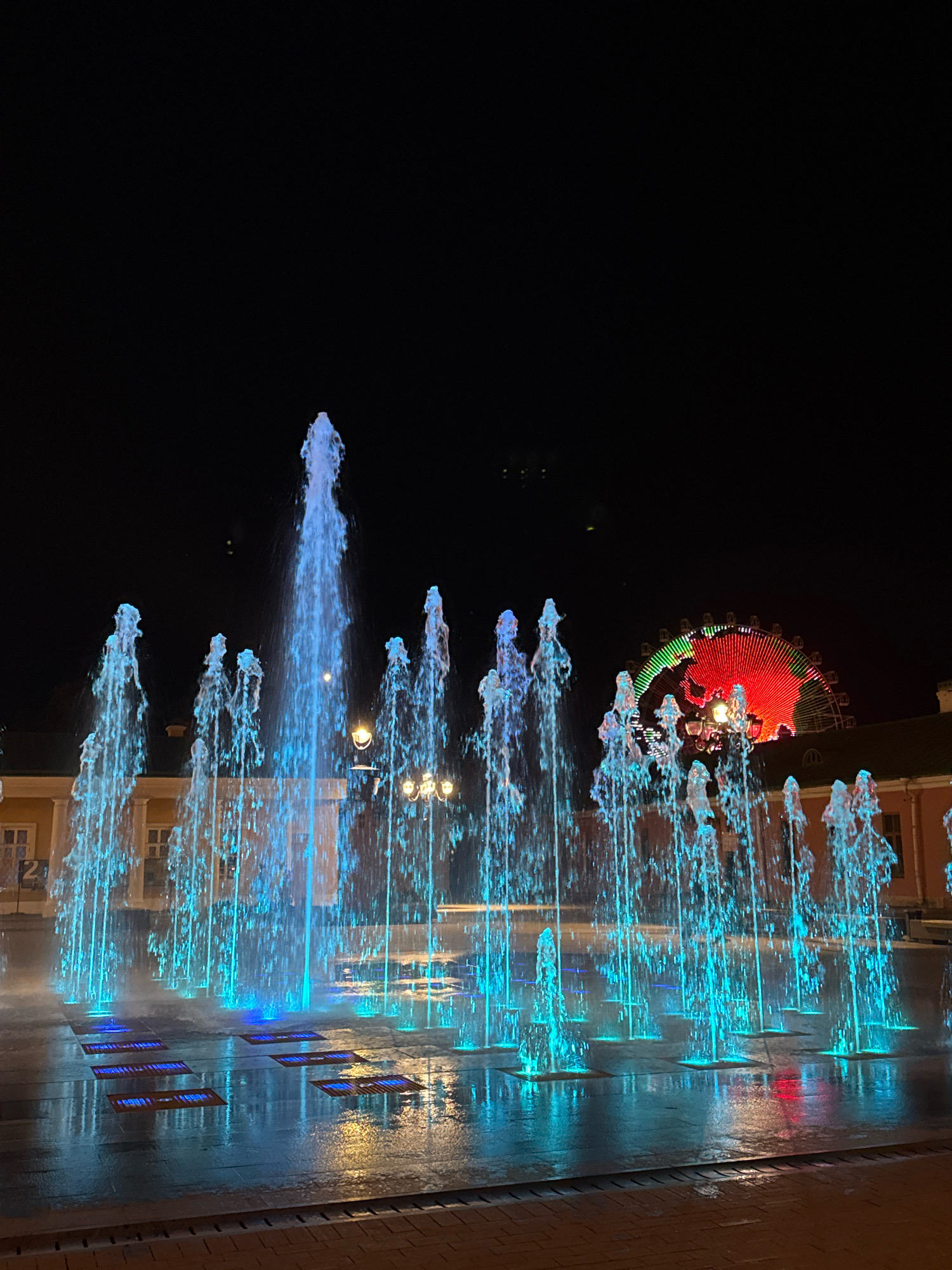 This image depicts a vibrant nighttime scene in Tver, Russia, likely at a public square or entertainment area. In the foreground, a large water fountain displays numerous jets of water illuminated with bright blue and turquoise lights. The water sprays upwards, creating a dynamic, almost ethereal effect against the dark sky. The ground around the fountain is wet and reflective, showing the illuminated water and the surrounding lights. In the mid-ground, there are buildings with arched windows and illuminated facades, suggesting a commercial or residential area. Further in the background, a Ferris wheel with colorful lights, predominantly red and green, is visible, indicating the presence of an amusement park or fairground. The overall atmosphere is lively and festive, with the combination of water features, colorful lights, and the iconic Ferris wheel creating a captivating urban spectacle.