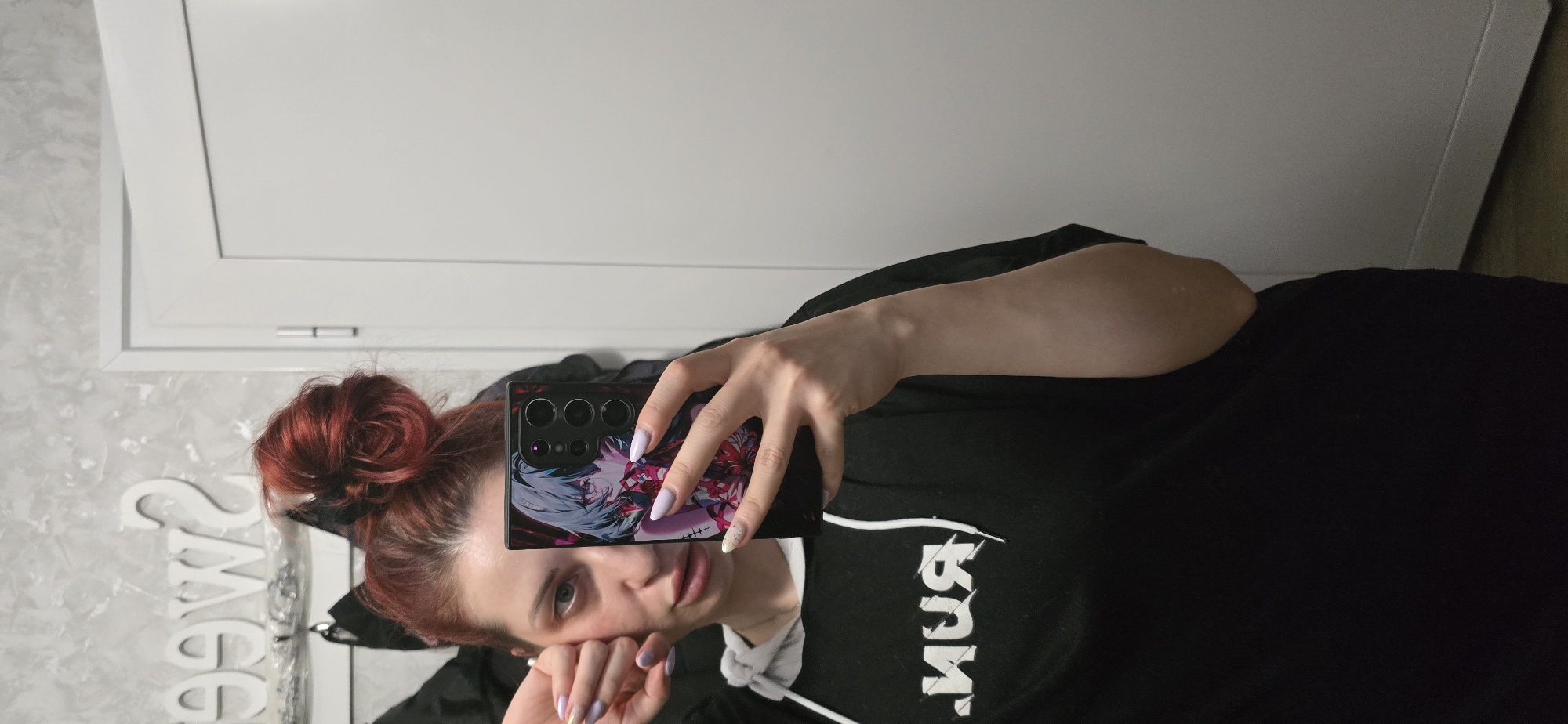 A young woman is captured in a selfie, holding a black smartphone with an anime-themed case featuring a character with white and red hair. Her left hand grips the phone, displaying light purple nail polish, while her right hand rests near her chin, showing a similar nail color. She has red hair styled in a bun and wears a black hoodie with a white drawstring. The hoodie has reversed white text on the chest, possibly "RUN" or a similar logo. Her expression is neutral, and she looks directly at the camera.
The setting is indoors, with a textured, light grey wall to her left, displaying partially visible, reversed white letters that spell "SWEET". To her right and in the upper background, there is a plain white door or cabinet. The lighting is even, suggesting a well-lit room. The image appears to be a casual self-portrait taken in a home environment in Burgas, Bulgaria.