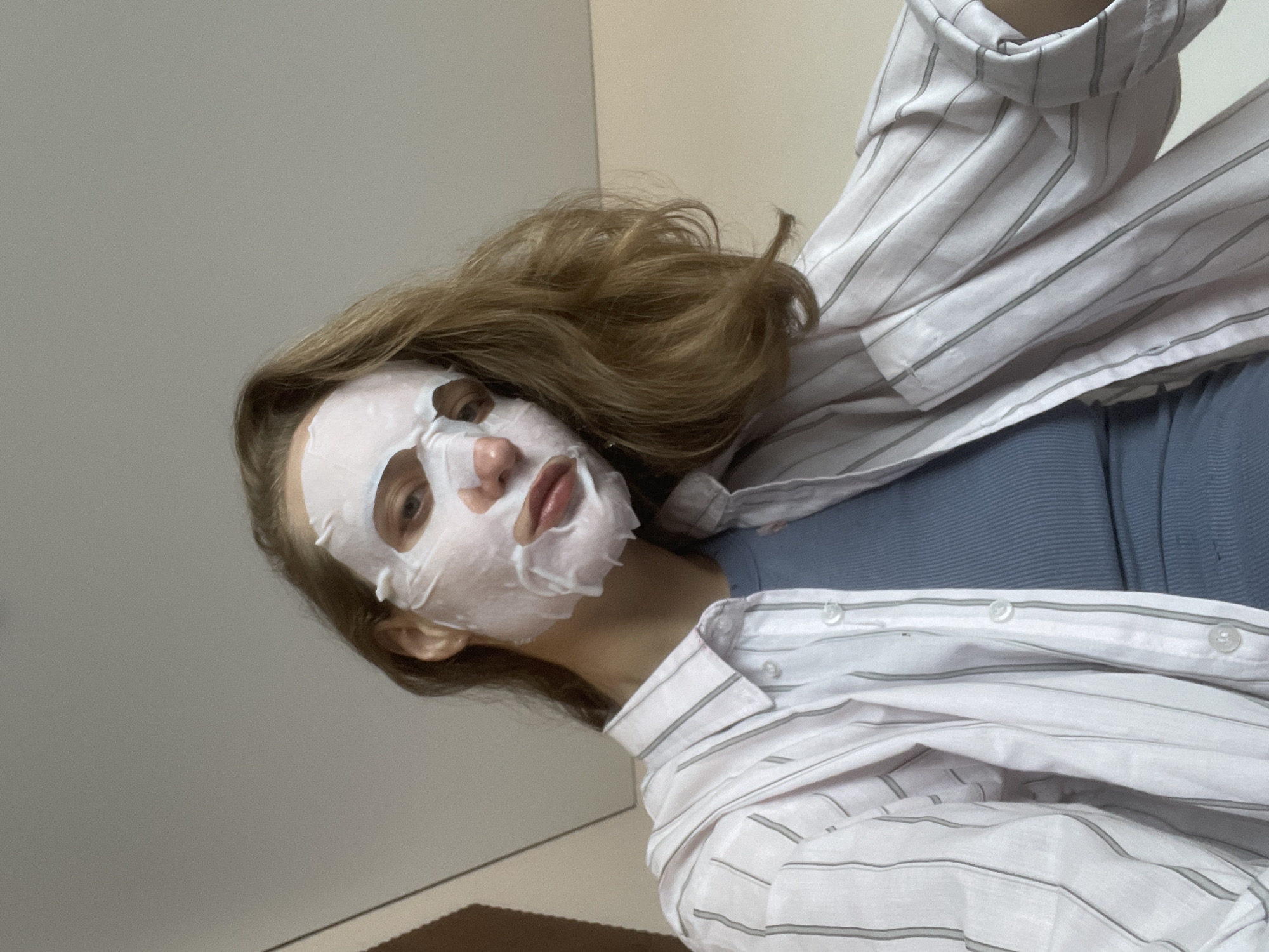 A young woman is reclining in an indoor setting. She is wearing a white facial sheet mask and a blue top under a white and grey striped shirt. Her light brown hair is styled in loose waves. The background is a plain wall. It is impossible to infer the location is Sepych, Russia from the image.