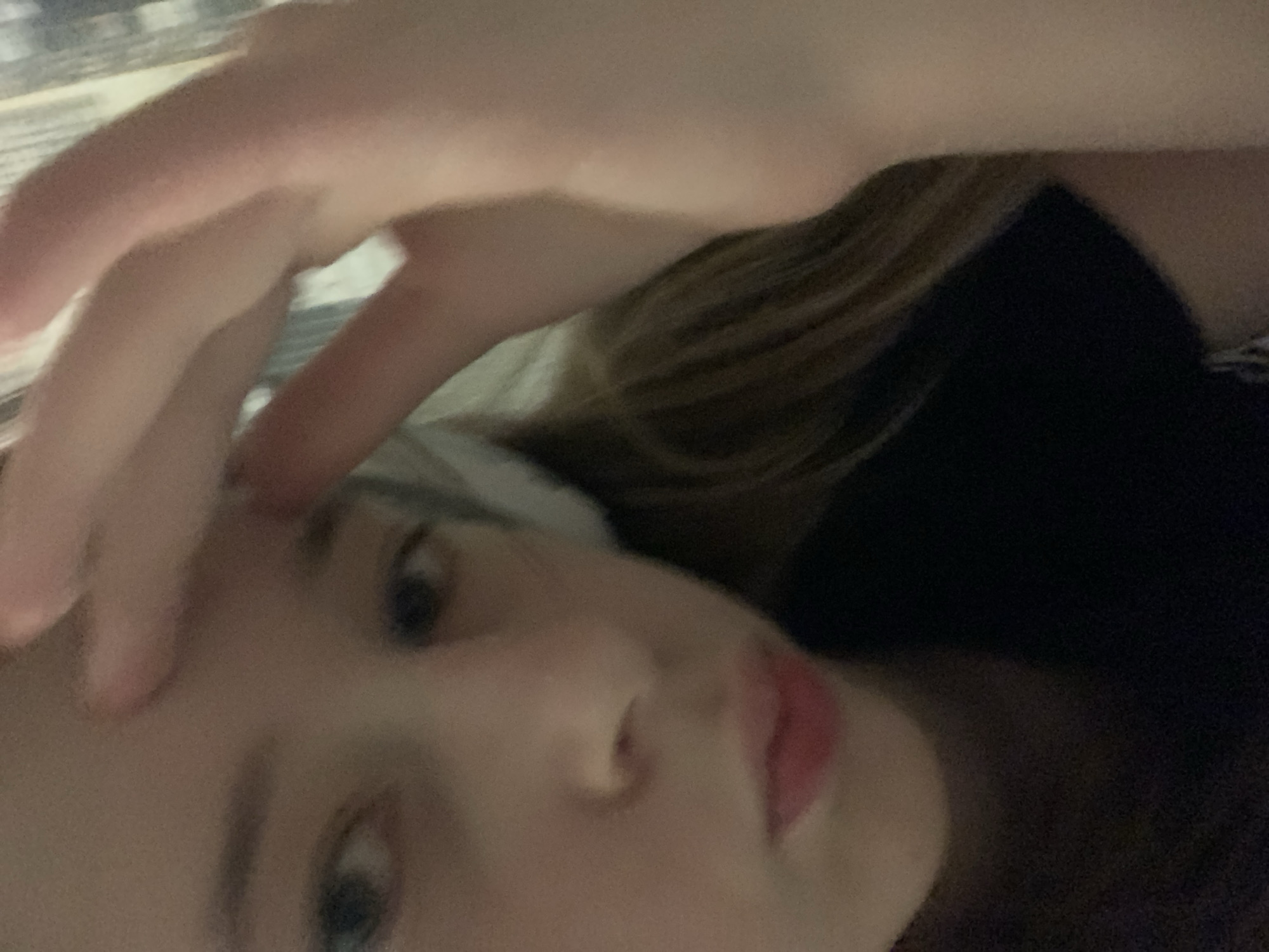 A person with fair skin is angled towards the camera, with their hand partially obscuring their forehead. They have blonde hair and are wearing a dark-colored garment. The background is out of focus.