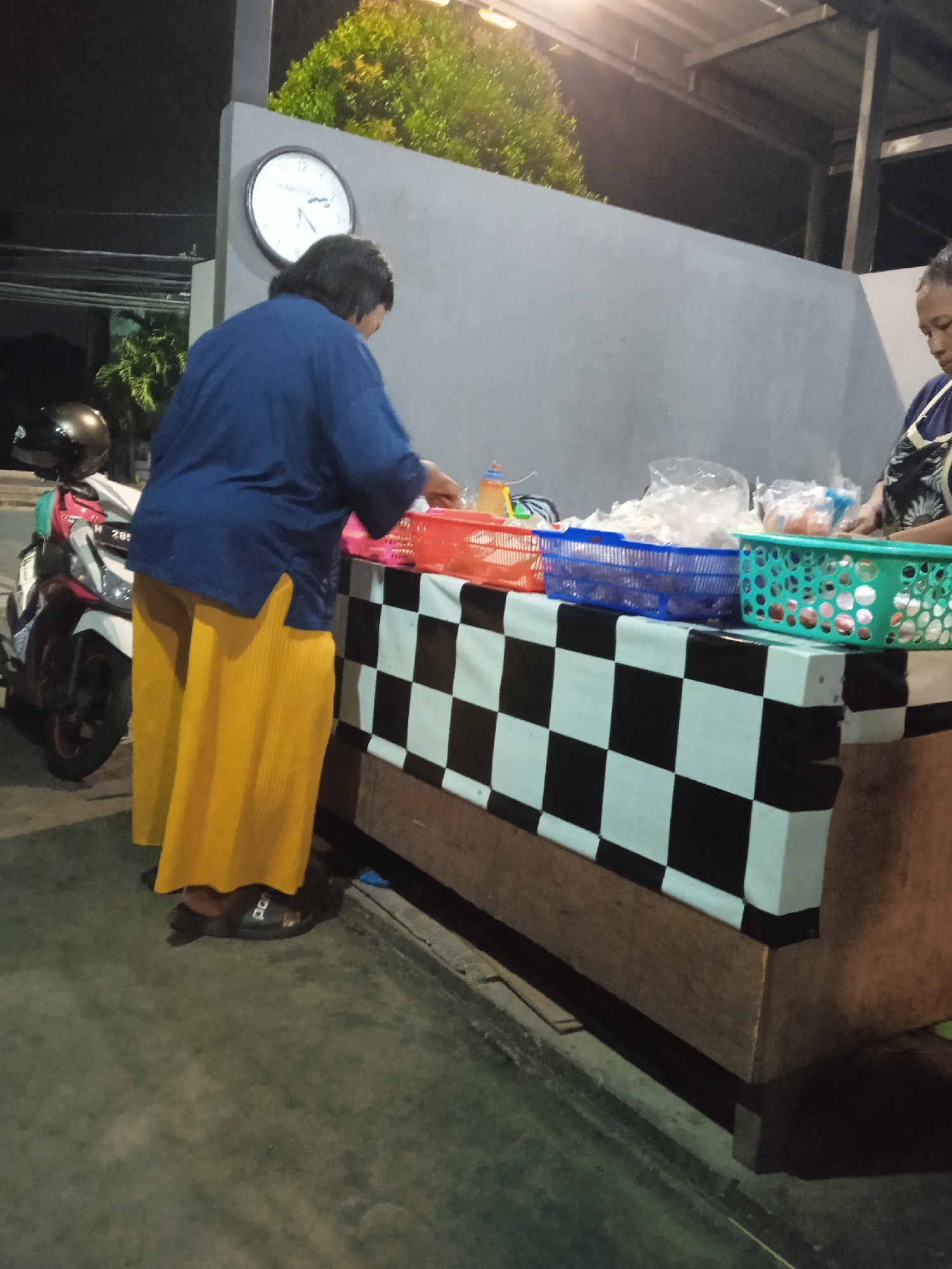 At night, on a sidewalk in Semarang, Indonesia, a person stands behind a wooden counter covered with a checkered black and white cloth. They are wearing a dark blue long-sleeved shirt and a bright yellow, pleated, floor-length skirt. Their feet are clad in black sandals. In front of them on the counter are several plastic baskets containing goods. To their left, a motorcycle is parked, its front wheel and fender visible. To the right of the counter, another person is partially visible, also appearing to be interacting with items on the counter. Behind the counter, a light gray wall is present, and a clock with Roman numerals hangs on it. Above the wall, green foliage is visible, suggesting the presence of plants or trees. The overall environment appears to be an outdoor stall or small shop.