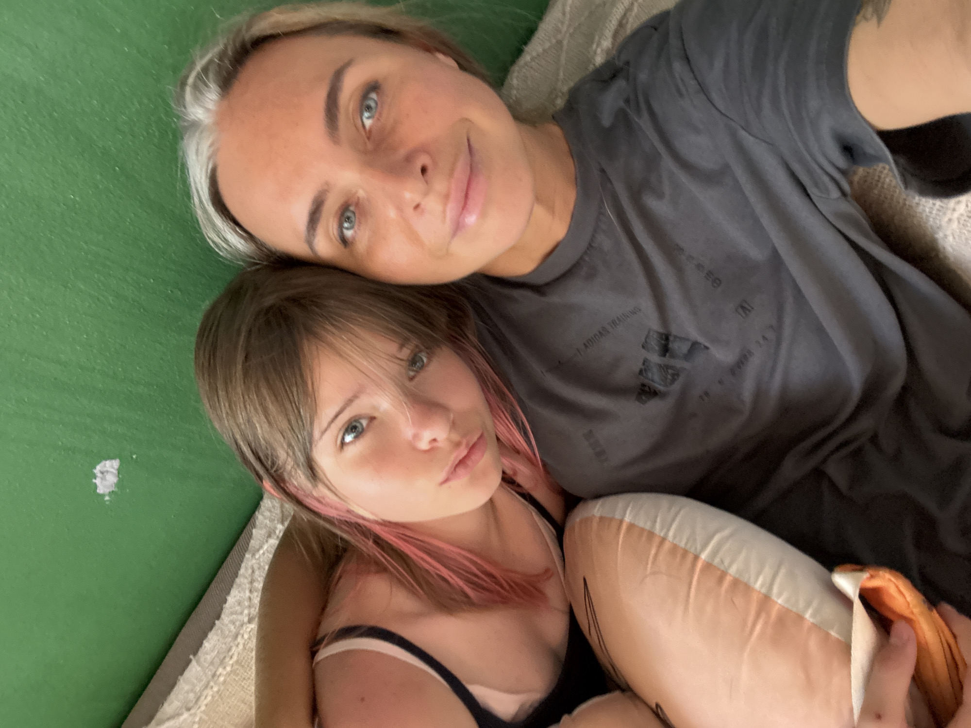 In Ko Samui District, Thailand, two individuals, an adult woman and a young girl, are captured in a close-up selfie. The woman, with blonde hair streaked with gray, has blue eyes and is wearing a dark gray t-shirt. The girl, with brown hair with pink highlights, also has blue eyes and is wearing a dark-colored tank top. They appear to be lying down, possibly on a bed, with a green wall visible in the background and a beige, textured surface underneath them. A pink and white pillow is positioned between them, partially obscuring the girl's body. The overall scene is intimate and casual, suggesting a personal moment shared between the two.