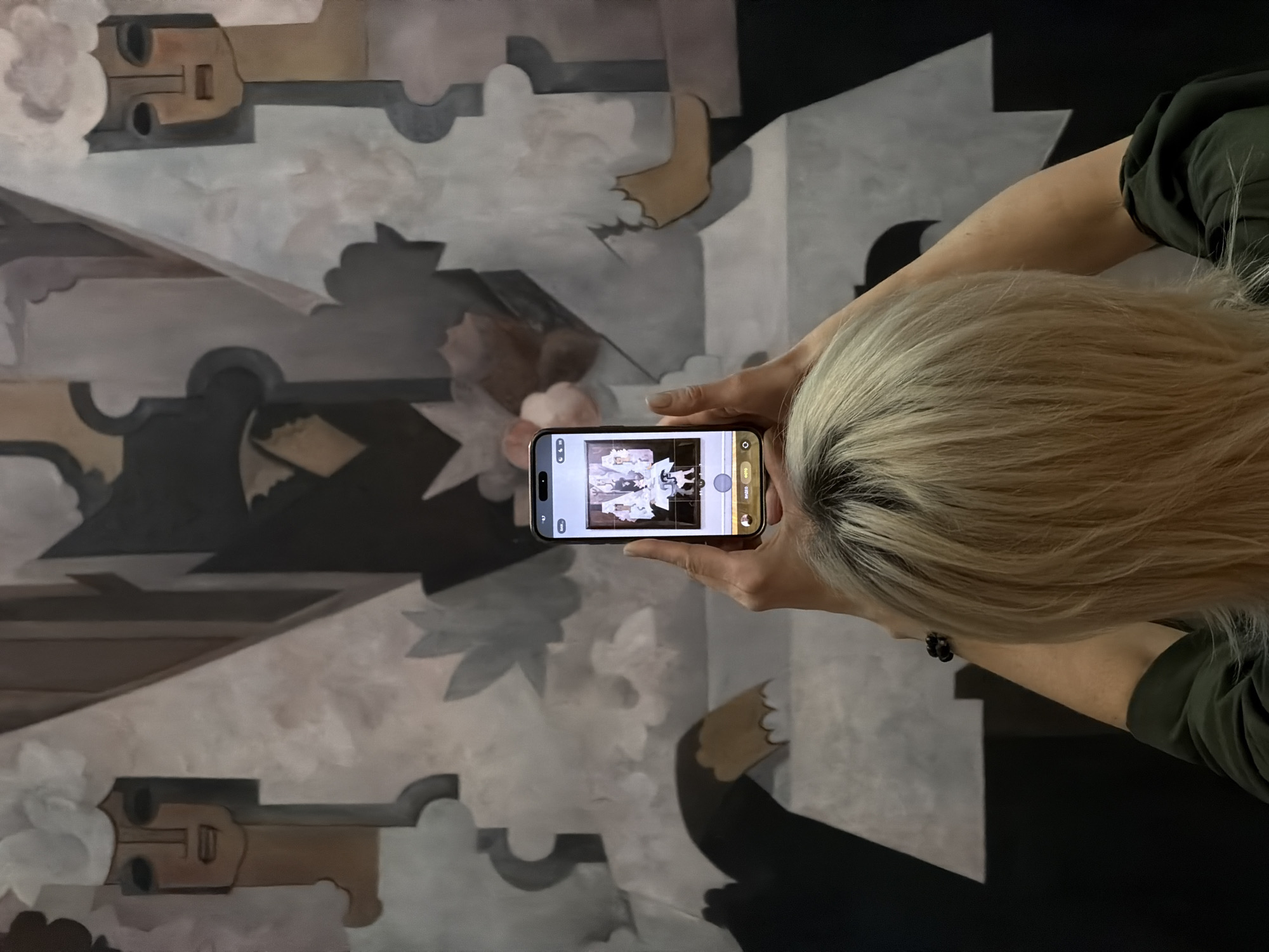 A woman with blonde hair and wearing a green long-sleeve shirt is holding a smartphone horizontally, taking a photo of a large abstract mural. The mural features geometric shapes and figures in shades of brown, gray, and white, with some elements resembling stylized faces or architectural forms. The scene is likely indoors, and the location is Malakhovka, Russia, based on contextual information.