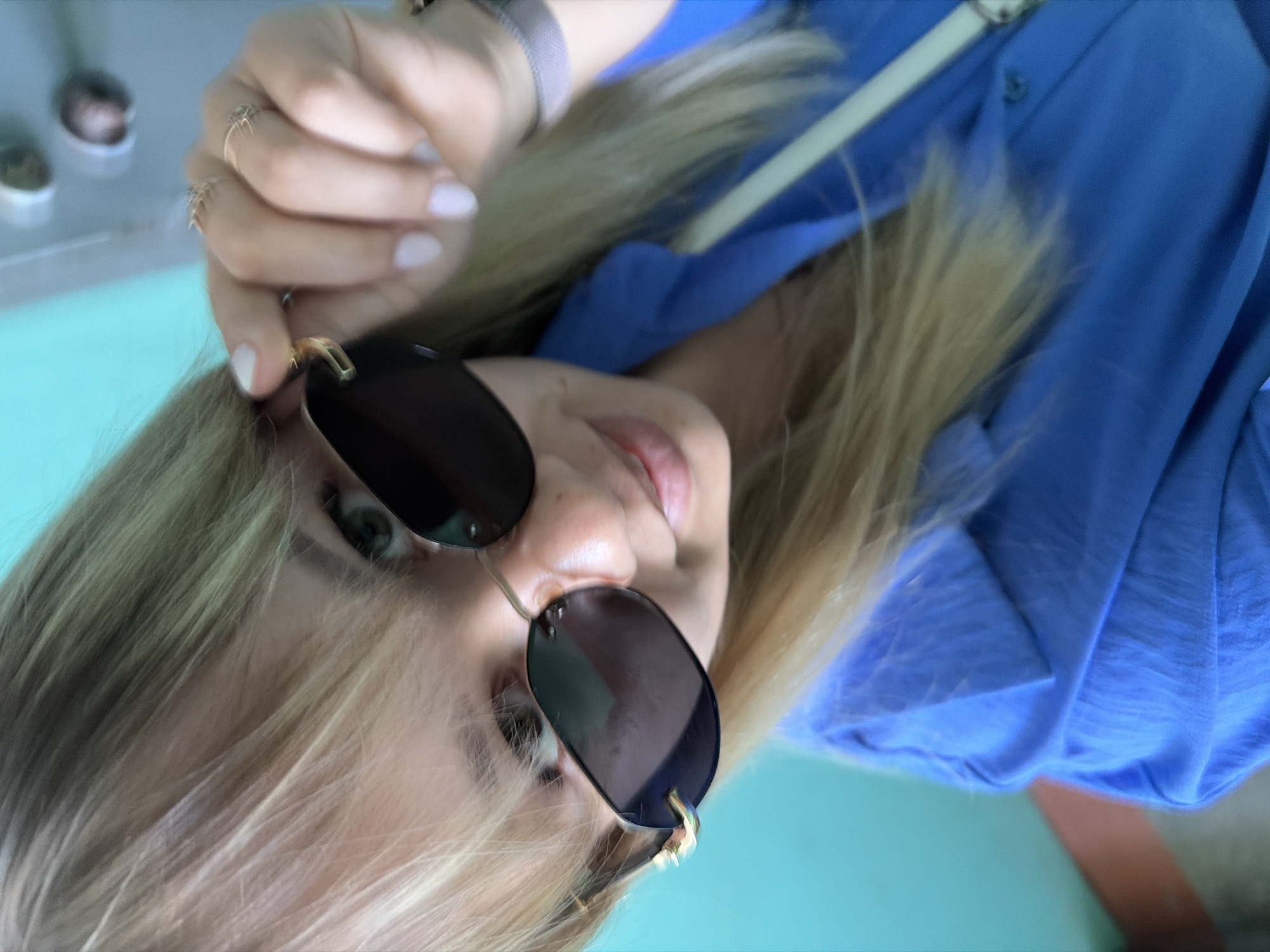 A blonde woman with multiple rings on her hand holds sunglasses in front of her face. She is wearing a blue button-down shirt and a light-colored purse strap is visible over her shoulder. The background appears to be a light blue or teal surface. The location is Chaikovsky, Russia.