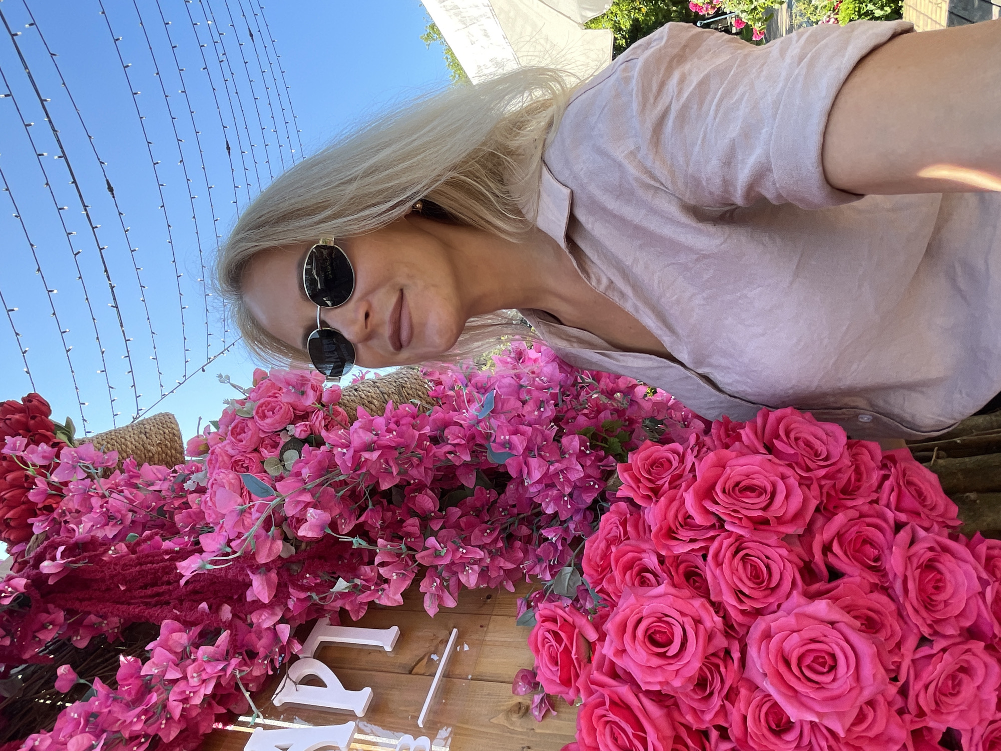 A blonde woman wearing sunglasses and a light pink shirt is standing in front of a vibrant display of pink and red flowers. She is looking to her right. The background features a clear blue sky with strings of lights overhead. The scene appears to be outdoors, possibly at an event or a decorative installation. There are wooden elements and a glimpse of white umbrellas and greenery to the right, suggesting an urban outdoor setting in Sochi, Russia.