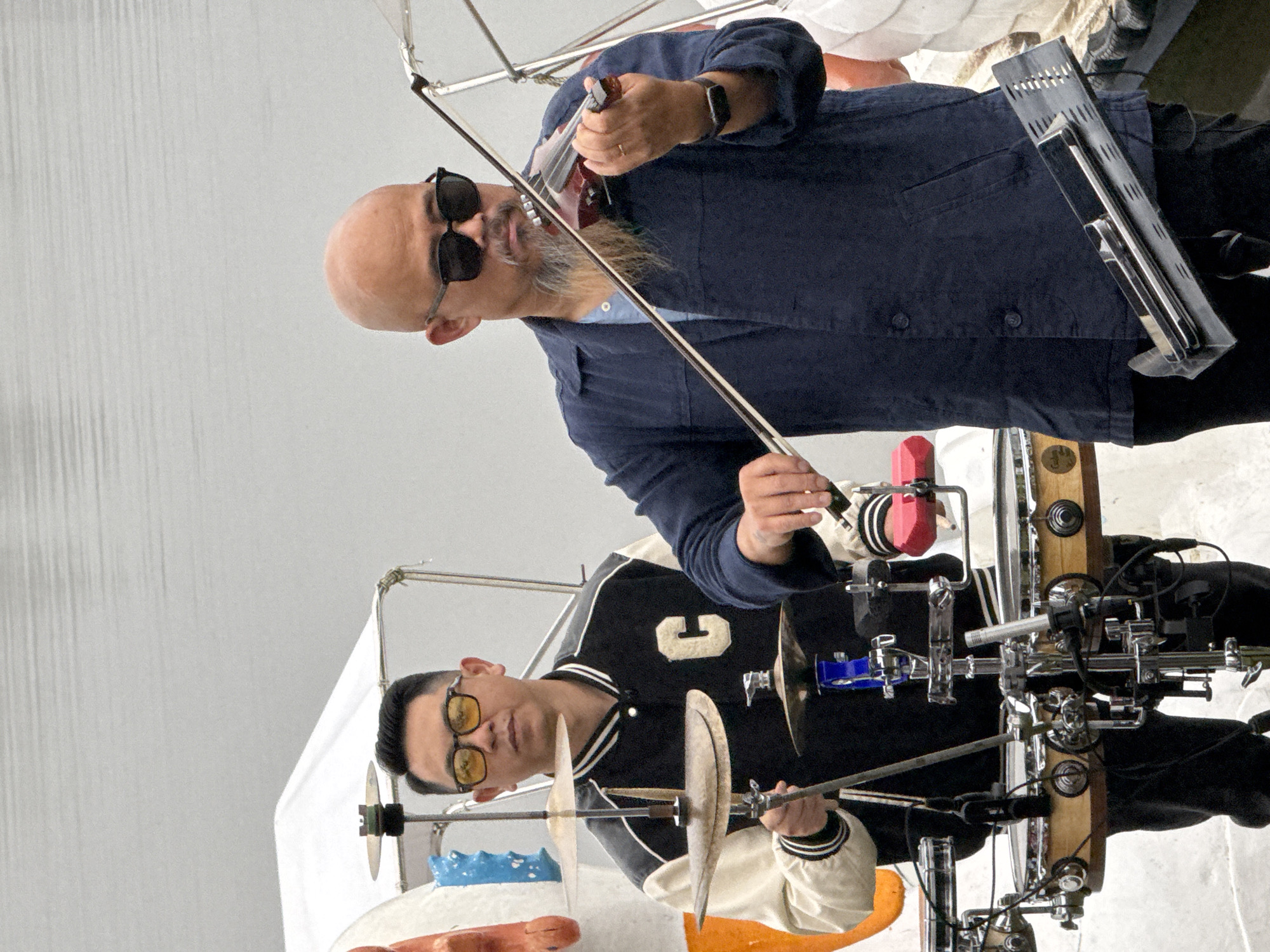 Two men are on a boat, performing music. The man on the left, wearing a black and white varsity jacket with a large 'C' on it and yellow-tinted glasses, is playing a drum set. The man on the right, bald with sunglasses and a goatee, is playing the violin. The boat appears to be on a body of water, possibly a lake or river, with a cloudy or overcast sky in the background. The overall scene suggests an outdoor performance, potentially in a tourist area given the boat setting. The image does not provide specific details to pinpoint the exact location within Da Lat, Vietnam.