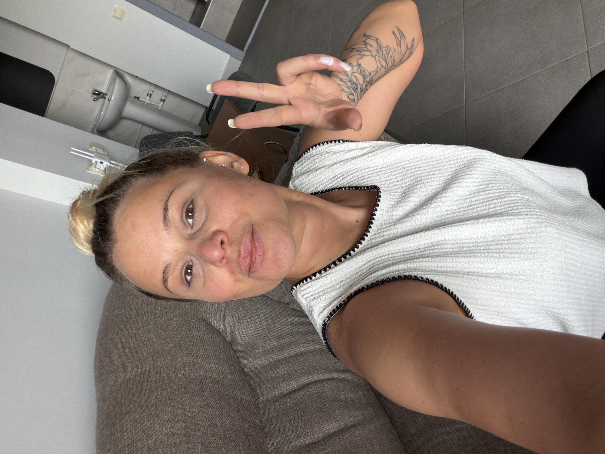 A woman with blonde hair in a bun is lying on a gray sofa, wearing a white top with black trim and black pants. She is holding up a hand with fingers extended in a "peace" or "victory" sign. A tattoo is visible on her upper arm. A sink and faucet are visible in the background. The location is not identified within the city of Sarapul, Russia.