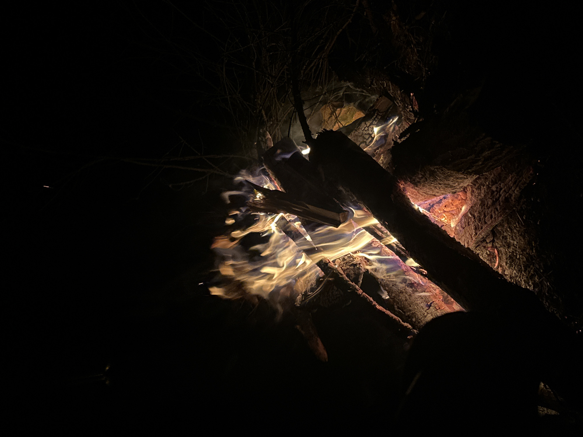 The image depicts a campfire burning in what appears to be a dark, outdoor setting. The focus is on the flames and embers of the fire, with wood logs arranged around and within the blaze. Some smoke is visible rising from the fire. The area surrounding the fire is dark, obscuring details of the environment, although hints of tree branches are visible in the background. The lighting is provided by the fire itself, creating a high contrast between the bright flames and the surrounding darkness.
The location is Папярнянскі сельскі Савет, Belarus. This is the only location information in this image.