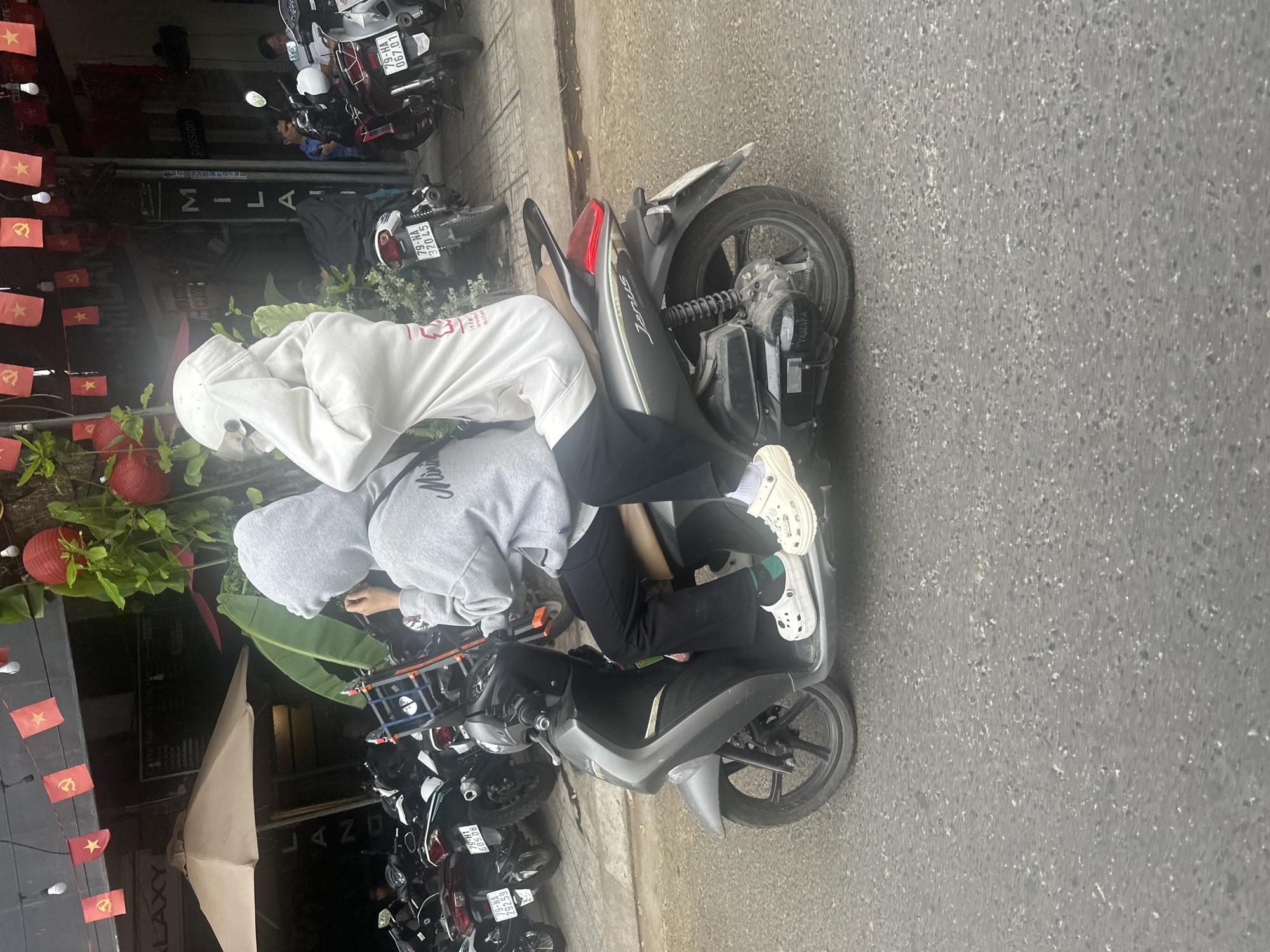 The image shows a motorcycle parked on the side of a street in Bac Nha Trang Ward, Vietnam. Two people are sitting on the motorcycle; one is wearing a gray hooded sweatshirt and black pants, and the other is wearing a white hooded sweatshirt and black pants. The motorcycle is parked near a row of other motorcycles and in front of a building with red flags and Vietnamese writing on it. The street is paved and there are some trees and plants in the background. The license plates of the motorcycles in the shot are shown, beginning with "79-."