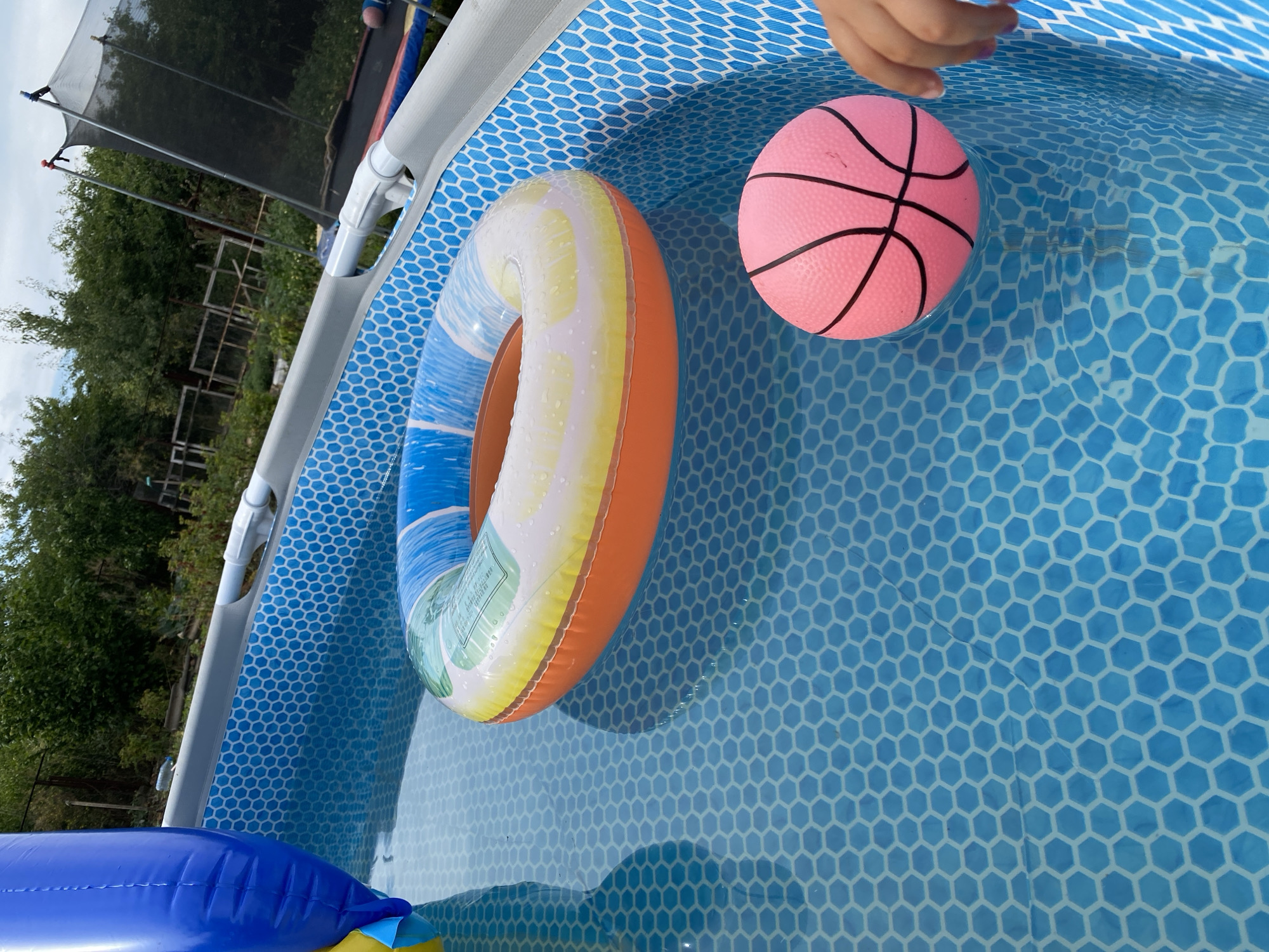 An overhead shot depicts a bright blue, hexagon-patterned inflatable swimming pool filled with clear water. A colorful inflatable ring, featuring bands of blue, white, yellow, and orange, floats in the center of the pool. To the right, a pink, miniature basketball floats, with a child's hand reaching towards it from the bottom right corner. In the background, a trampoline with a black safety net is visible, along with lush green trees and a partially visible blue inflatable toy. The scene suggests a recreational setting, likely a backyard in Kropyvnytskyi, Ukraine.