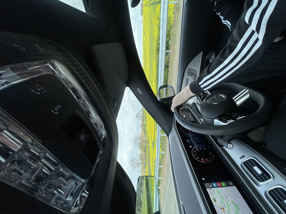 The image shows the interior of a car, from the driver's perspective. The driver is holding the steering wheel with their right hand and their left arm is resting on the seat. The driver is wearing a black jacket with white stripes. The dashboard shows a digital display with a speedometer, a temperature gauge, and a navigation system. The car is traveling on a road with a yellow field on the right side. There is a metal fence separating the road from the field. The car is driving towards a row of trees in the distance. The GPS shows a location that is in close proximity to the city of Woburn, United Kingdom, which is located at a latitude of 51.99 and a longitude of -0.618. The coordinates provided indicate a rural setting, likely in a field area outside of the main town center. The sky is cloudy. The car's interior is illuminated by natural light. The overhead light is off. The driver's side window is down, and the interior mirror is tilted. The dashboard is displaying a number of 37. The rearview mirror is visible. The steering wheel is leather-wrapped with a silver emblem. The buttons on the overhead console indicate an SOS function, a microphone, and a button labelled 'SOS'. The door handle has a button. The seat is covered in a dark fabric. The car has a black interior.