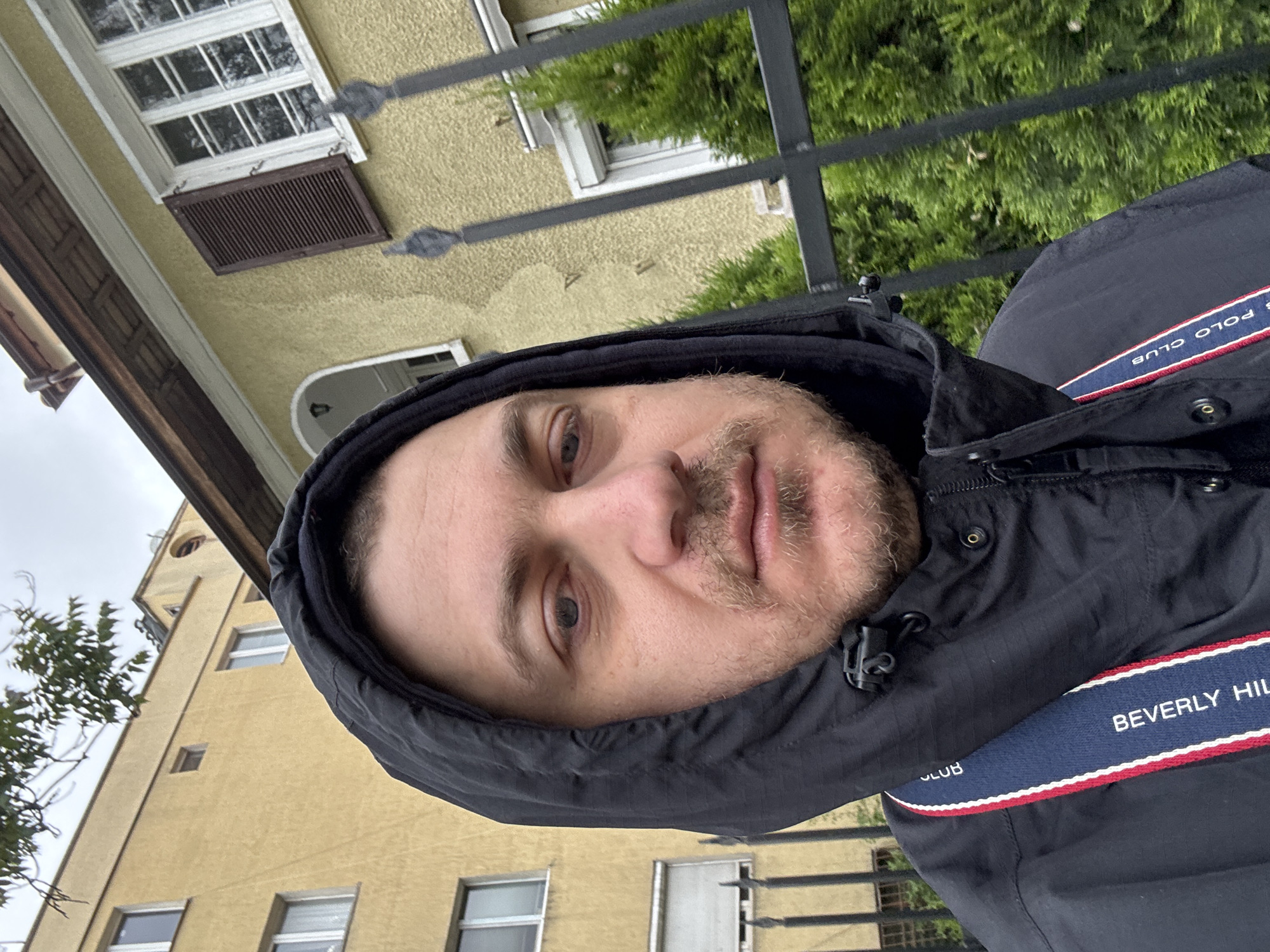 A selfie shows a man wearing a black hooded jacket standing outside in Sofia, Bulgaria. The man has light skin, a short beard, and is looking directly at the camera. His jacket has a stripe with the words "BEVERLY HILLS POLO CLUB" visible. In the background are buildings with yellow facades and white-framed windows, partially obscured by a wrought iron fence and some green foliage. The sky appears overcast.