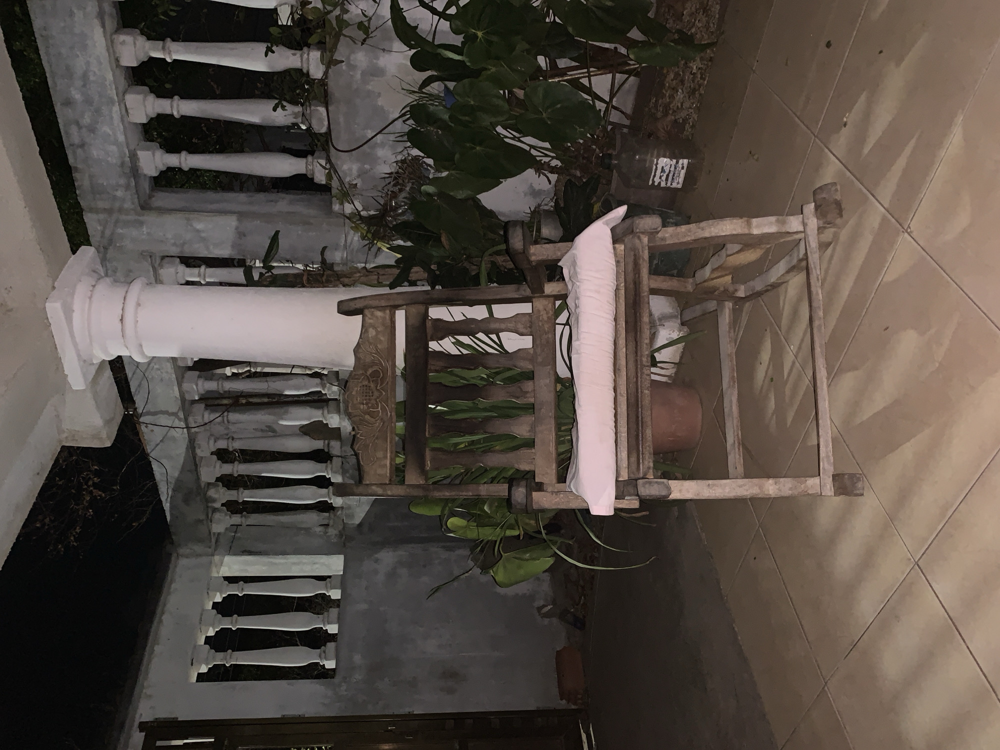 The image shows a patio setting in Santa Ana, Venezuela. A weathered, wooden chair stands on tiled flooring. A white cushion is draped over the chair. The chair is surrounded by lush greenery, including potted plants. The backdrop includes a building with a white column, decorative balustrades, and a window with wrought iron bars. The lighting suggests it's evening or a dimly lit outdoor space.