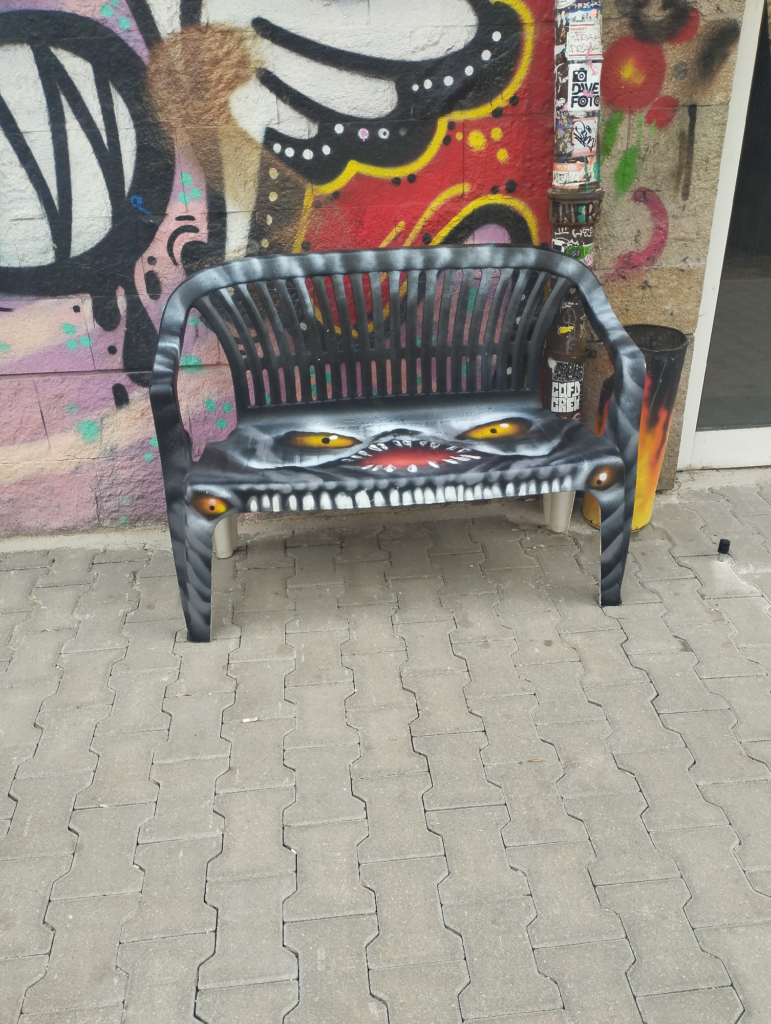 The image shows a black plastic bench painted with a monstrous face on a sidewalk next to a wall covered in graffiti. The bench has yellow eyes, a large mouth with sharp white teeth, and red lips. The graffiti on the wall includes abstract shapes, lines, and colors such as black, white, red, and green. A metal pipe with stickers is visible behind the bench, near the wall. A trash can is next to the bench on the right. The sidewalk is made of gray interlocking paving stones. The location is in Sofia, Bulgaria.