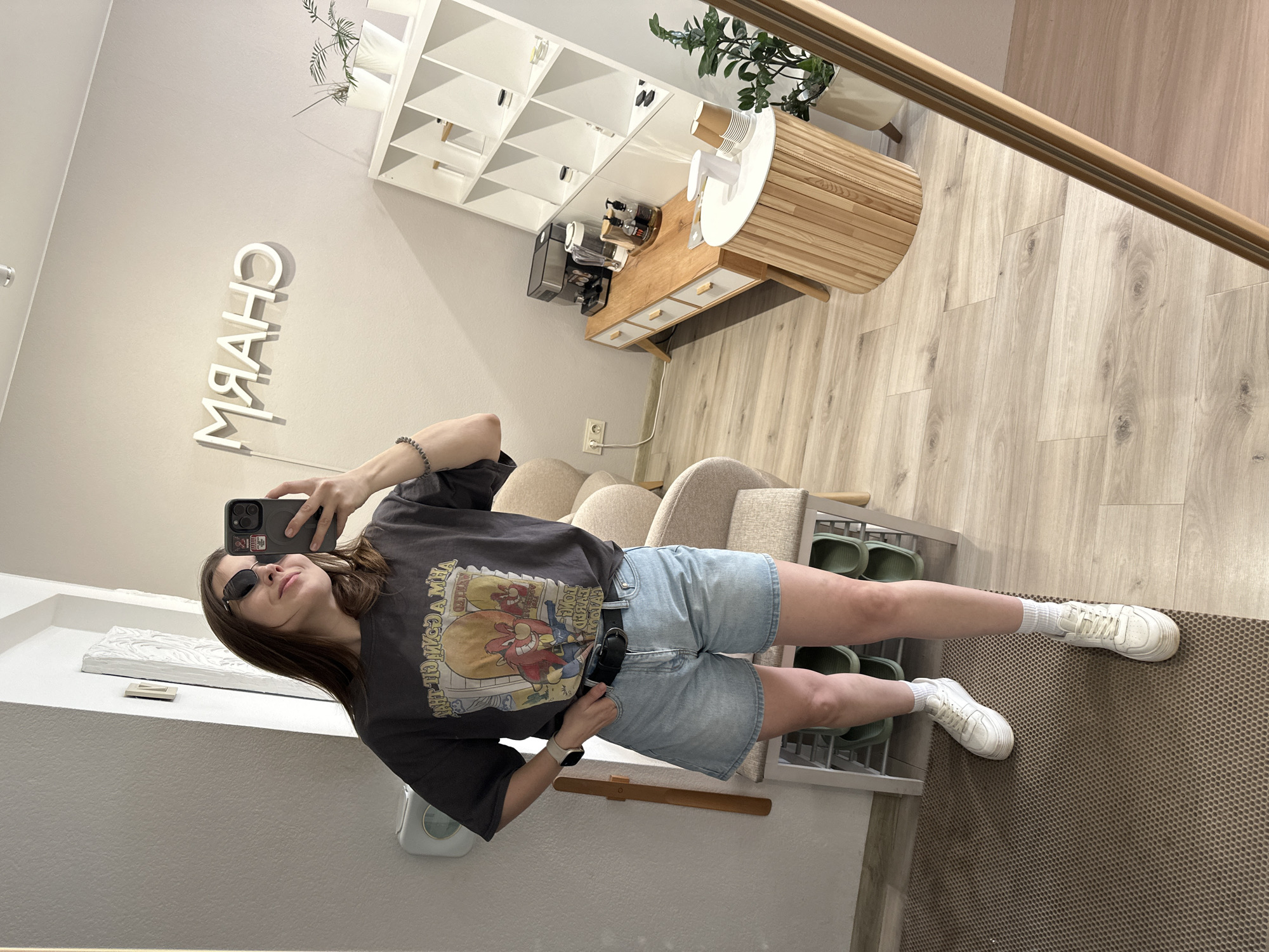 A young woman stands in front of a mirror, taking a selfie. She is wearing sunglasses, a grey graphic t-shirt, denim shorts, and white sneakers. The reflection shows a brightly lit interior with modern furnishings: white shelving units, a counter with coffee-making equipment, and a chair. A neon sign reading "CHARM" (reversed in the mirror) is visible on the wall. The floor is covered with a patterned rug. The environment suggests a retail or service establishment. The location is Perm, Russia, as indicated by the neon sign.