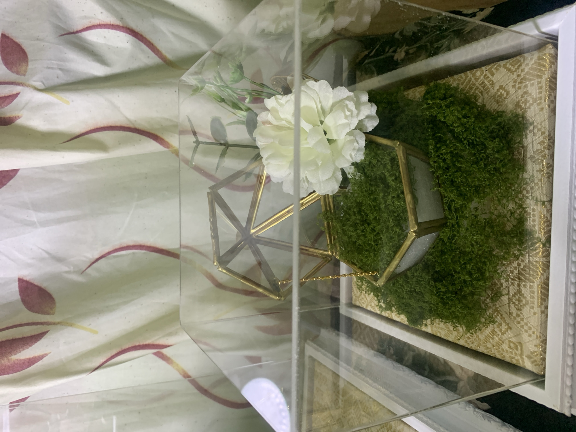 The image depicts a decorative display case, likely for a special occasion, containing a geometric gold-toned frame with a white flower and green moss. The case itself is clear, revealing the objects inside. The background is a cream-colored curtain with a subtle red and pink floral pattern. The objects are arranged on a decorative fabric that has a gold and white pattern. The display is placed on a white base with decorative molding. No specific location in Kuala Pembuang, Indonesia, can be determined from the image.