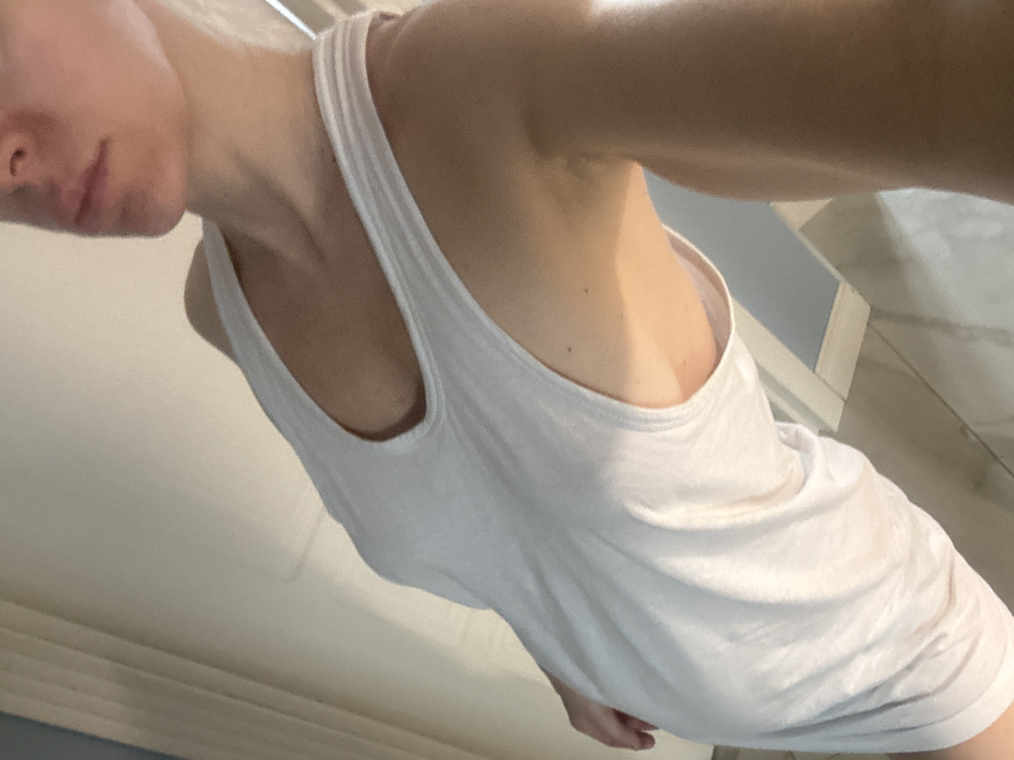 A close-up, slightly angled, interior shot depicts a person wearing a white tank top. The visible portion of the person includes part of their face, neck, shoulder, and arm. The tank top features wide straps and a scoop neckline. The background shows elements of a room with light-colored walls and a tiled floor. A piece of furniture or a fixture with a light blue surface is visible. The location is unknown.