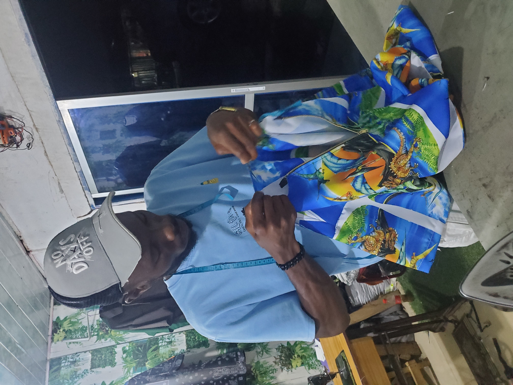 A man wearing a light blue t-shirt and a grey baseball cap is shown in what appears to be a tailoring shop. He is holding a garment with a vibrant blue fabric featuring a graphic print of dragons. The man is looking down at the garment, and his hands are positioned as if he is working on it. A blue measuring tape is draped around his neck. In the background, a sewing machine is visible on a wooden table to the left. The environment seems to be indoors, with a window or door on the right side, revealing a dark exterior. The overall impression is that of a working tailor's studio, possibly in the city of Jos, Nigeria, given the context provided in the prompt, though this is not visually confirmed by the image itself.