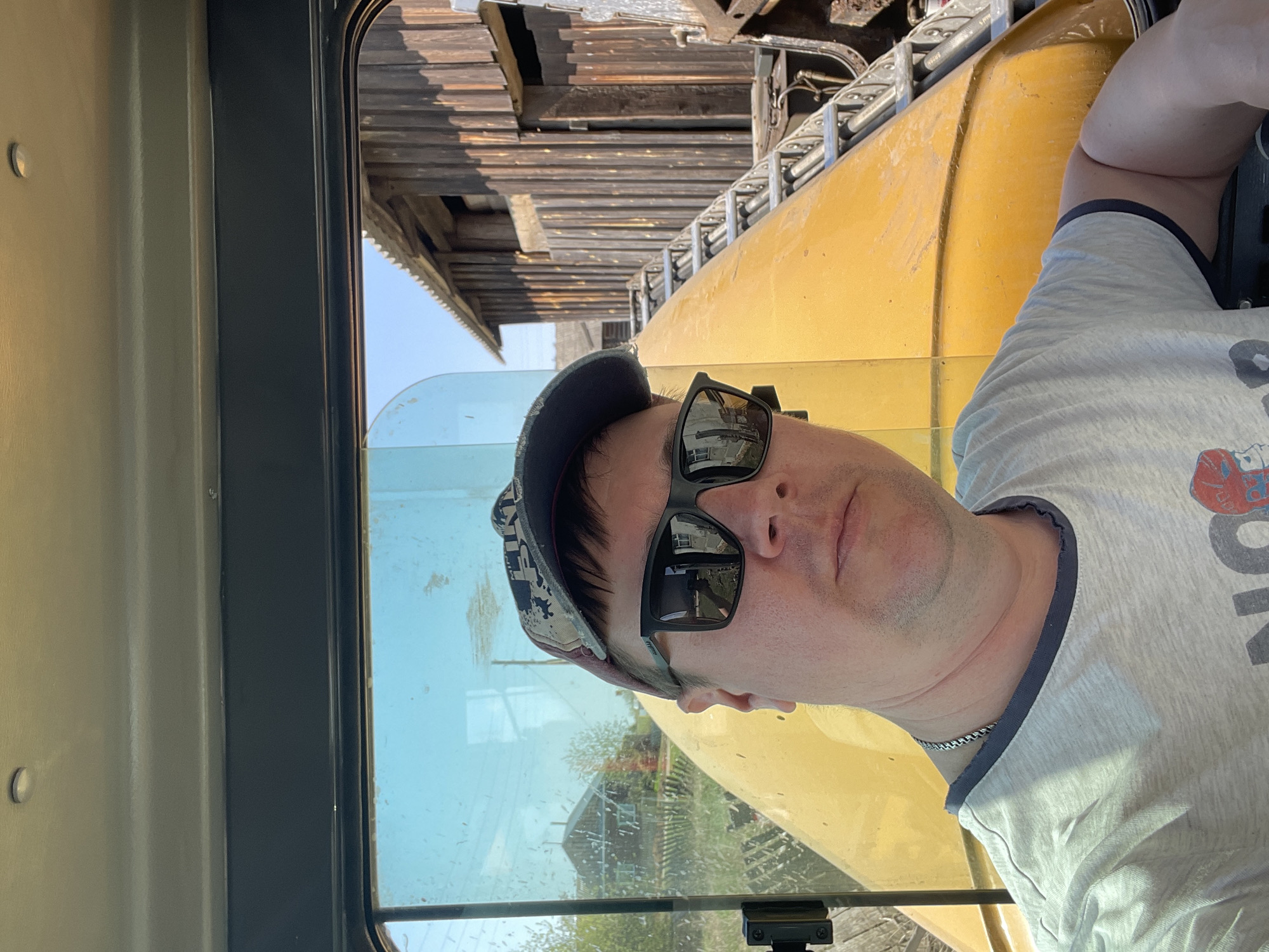 A man wearing a baseball cap and sunglasses is pictured inside the cab of a yellow construction vehicle in Поедуги, Russia. The man is wearing a gray t-shirt and a silver chain. Outside the window of the cab, there is a partially visible wooden building, and there is equipment visible above the man.