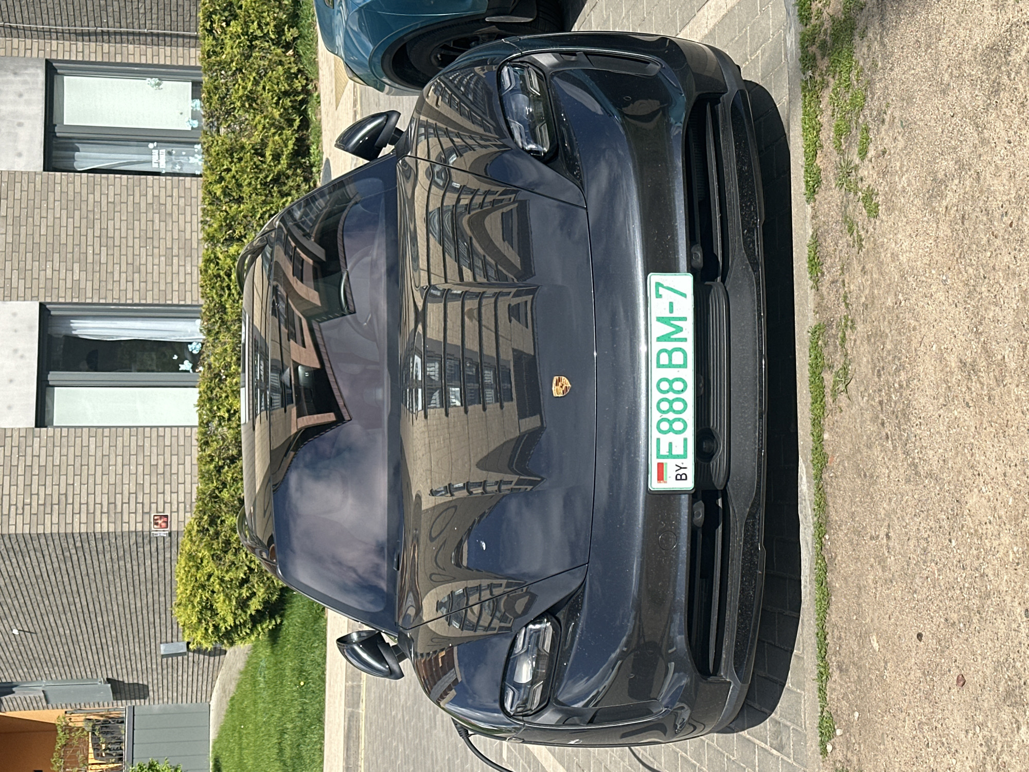 A black Porsche Taycan is parked on a paved surface in Бараўлянскі сельскі Савет, Belarus. The car has a Belarusian license plate that reads "E888 BM-7". The reflection of a building is visible on the car's windshield. Nearby is a building with brick walls and windows. Adjacent to the car is a strip of grass. Another car, partially visible, is parked behind the Porsche.